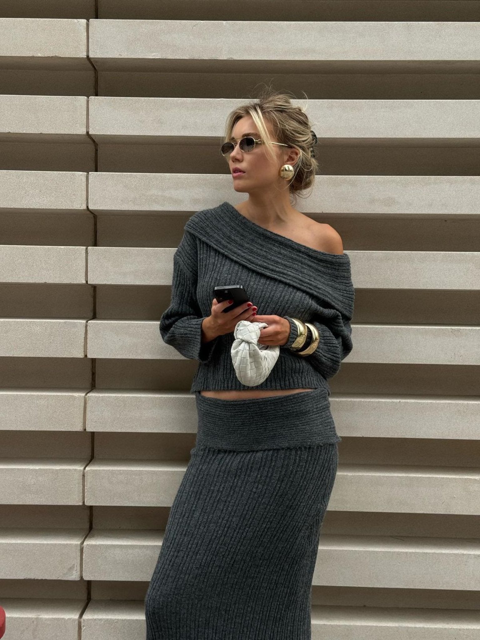 A person wearing the Ayla set in gray, featuring an off-shoulder knitwear top paired with a matching maxi skirt, leans against a ribbed concrete wall. They are holding a phone and a white bag, adorned with sunglasses and large hoop earrings, while gazing to the side.