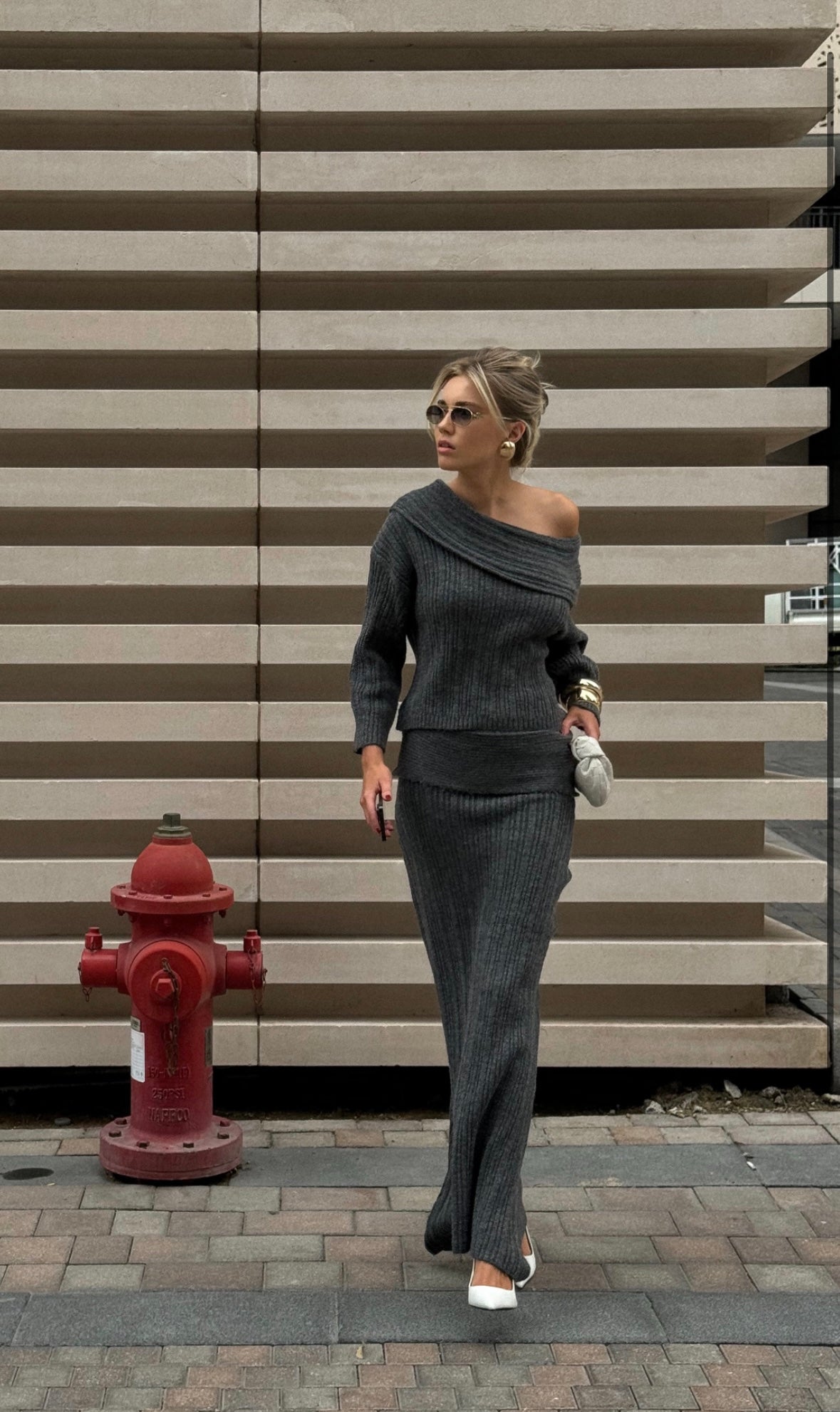 Dressed in a stylish Ayla set featuring an off-the-shoulder gray knit top paired with a maxi skirt, the person, accessorized with sunglasses and carrying a white clutch, walks along a brick sidewalk. A red fire hydrant stands next to horizontal beige panels in the background.
