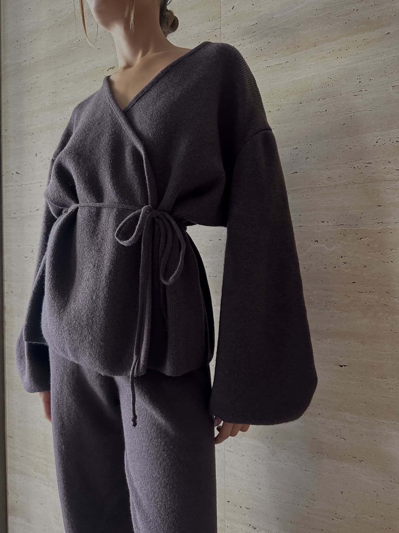 Clad in the Azalia Set, a loose dark gray wrap sweater with a tied waist and wide-leg trousers, the person embodies minimalist elegance. Against a textured off-white wall, the image captures the clothing's flow and partial profile.