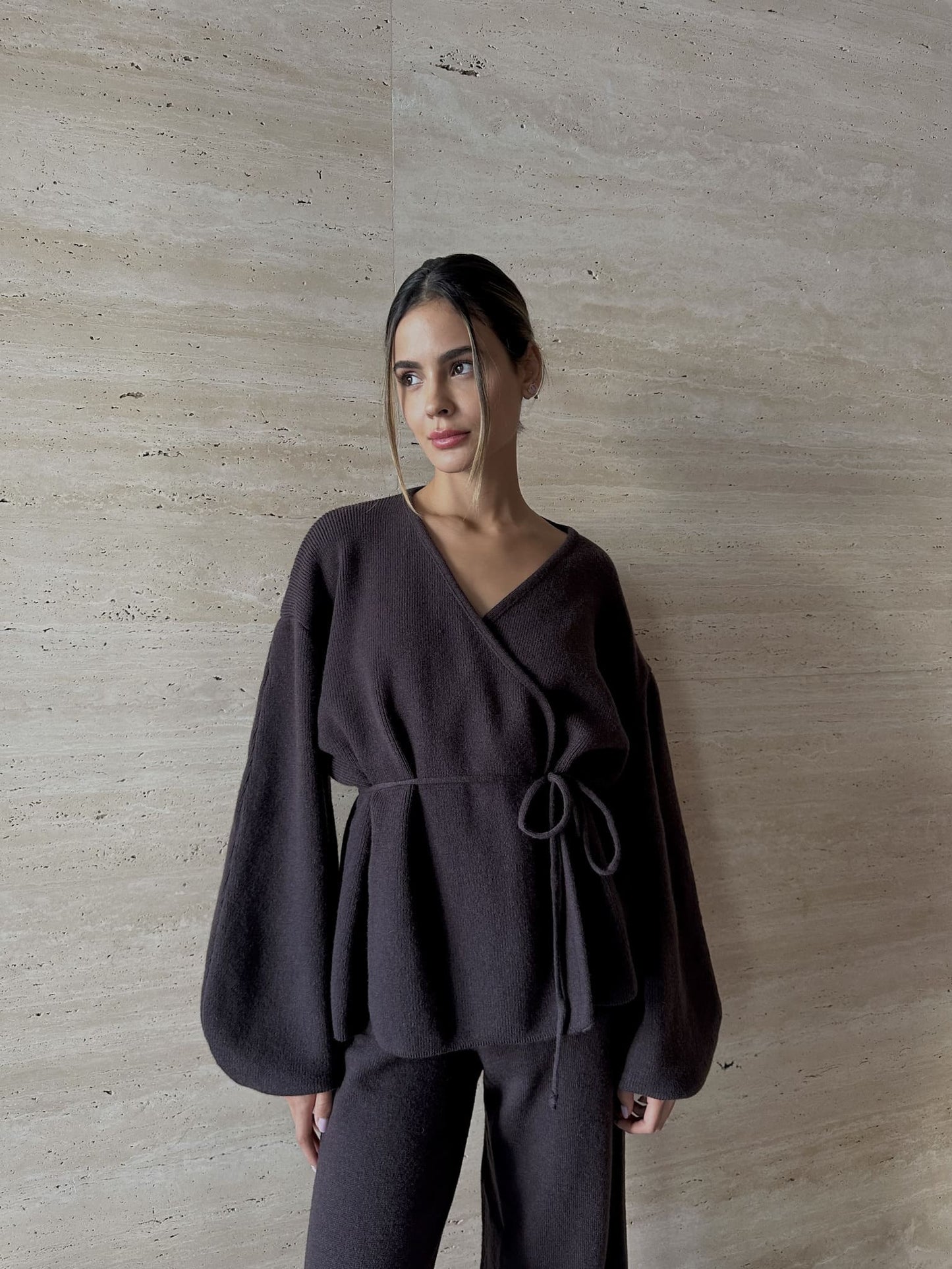 A woman models the Azalia Set, consisting of a black belted wrap top with voluminous sleeves and wide-leg trousers. She stands against a light stone wall, gazing sideways with a neutral look. Her pulled-back hair highlights her features with minimalist elegance.