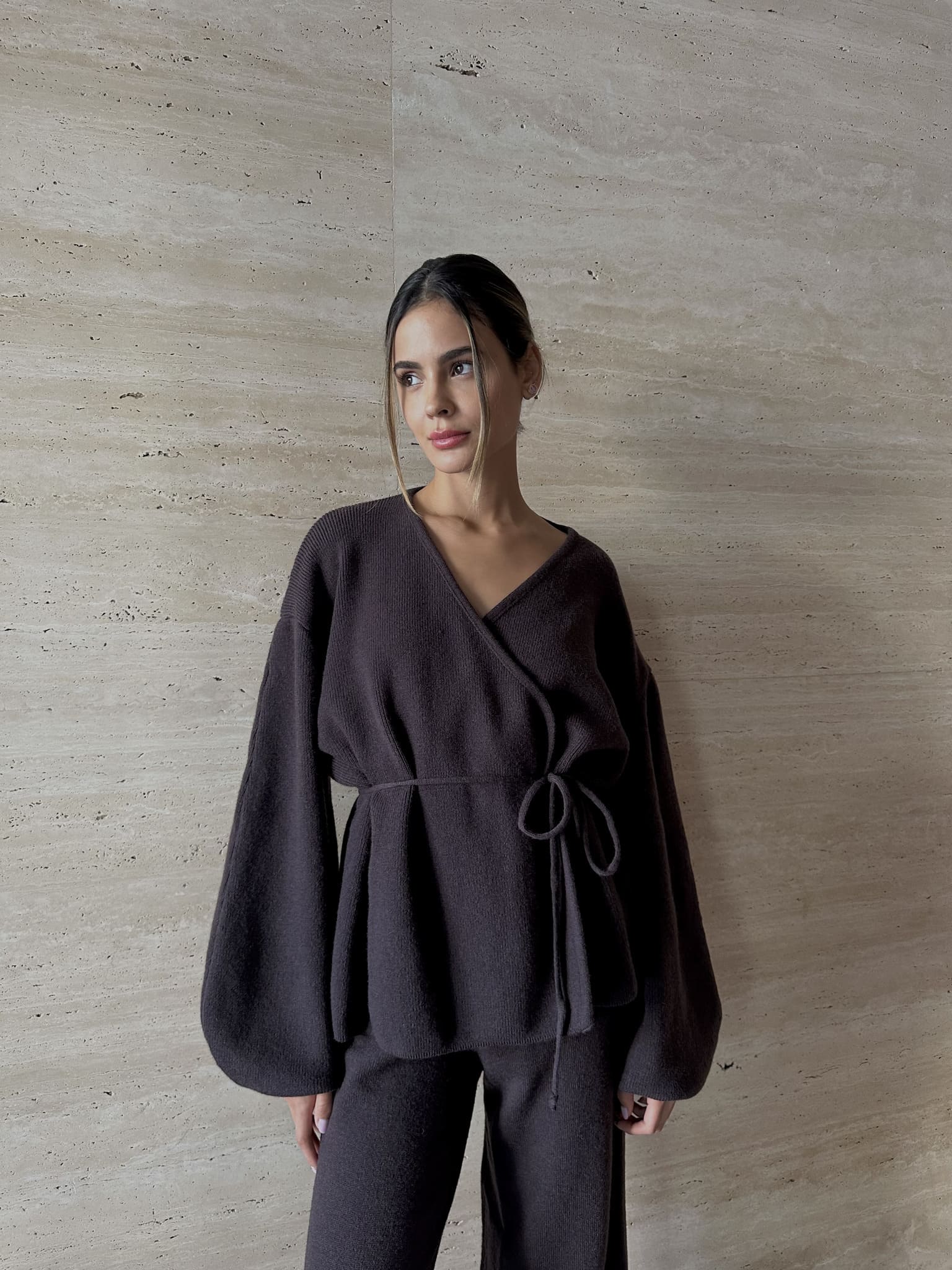 A woman models the Azalia Set, consisting of a black belted wrap top with voluminous sleeves and wide-leg trousers. She stands against a light stone wall, gazing sideways with a neutral look. Her pulled-back hair highlights her features with minimalist elegance.