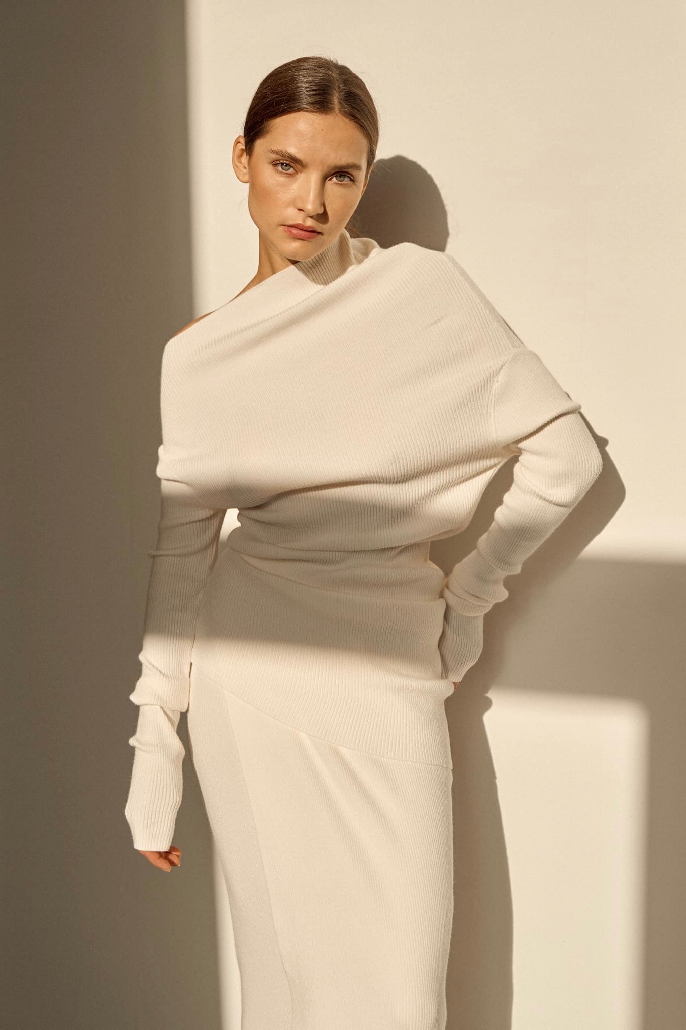Against a beige wall, a person is dressed in an Avani Set featuring an off-shoulder, long-sleeved cream ensemble. The asymmetrical draped neckline lends a touch of elegance as they pose with their hands on their hips, partially lit by sunlight that creates complex shadows on the wall.