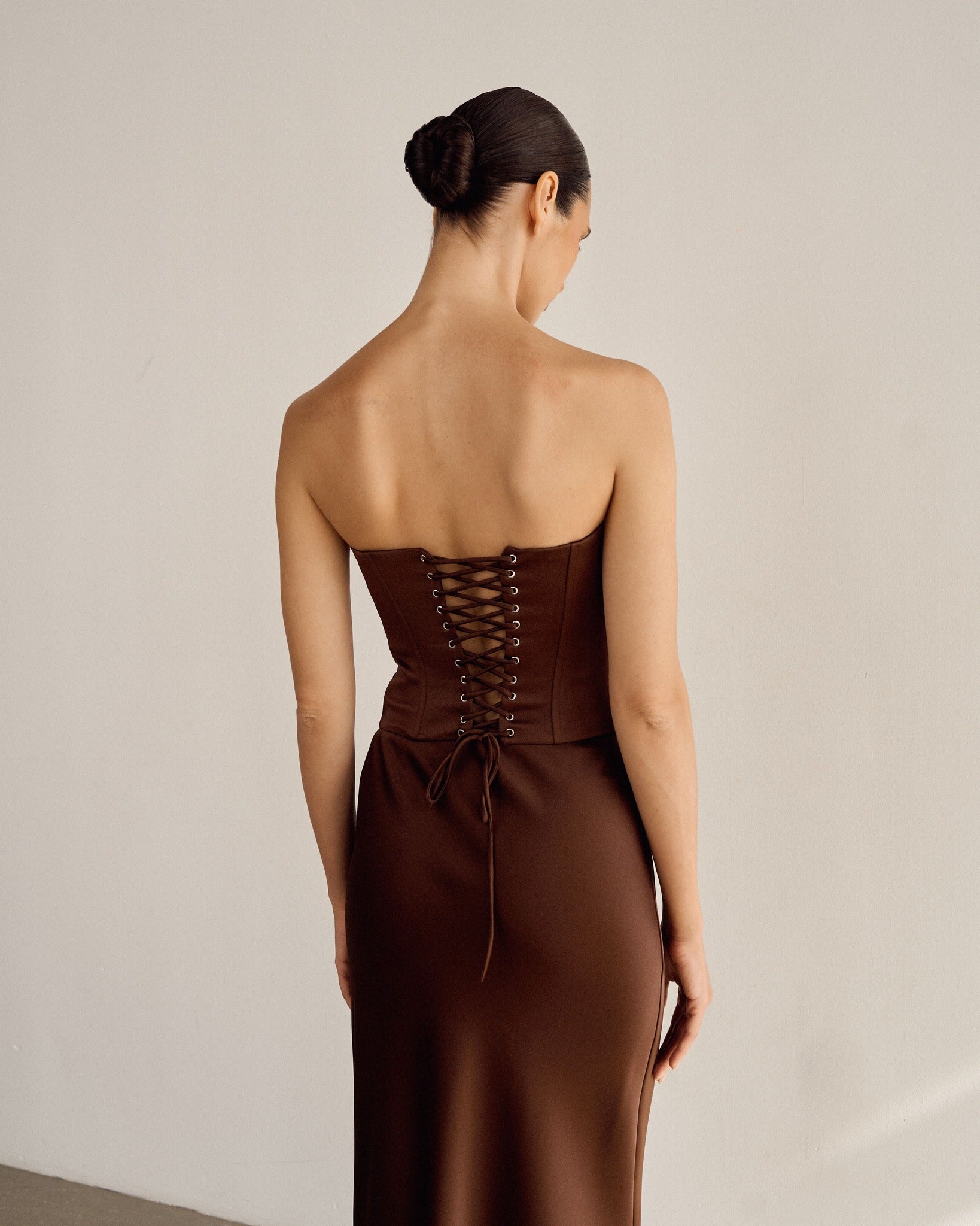 A woman with dark hair in a bun wears the Regina corset top, crafted from satin fabric in a strapless brown design with a lace-up back, standing against a plain white wall. The garment extends gracefully to the floor, capturing the elegance of Regency fantasy in her attire.