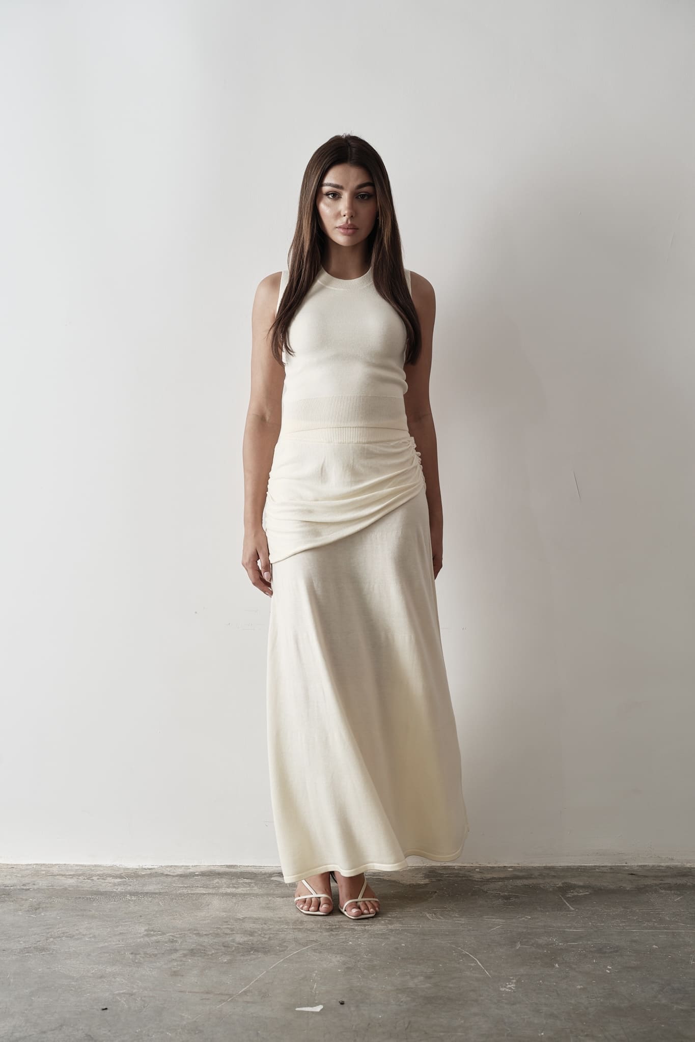 Model wearing a white Andrea Set featuring a sleeveless knit top with gathered detailing and a matching maxi skirt, styled with white strappy sandals, posing against a grey and white textured background