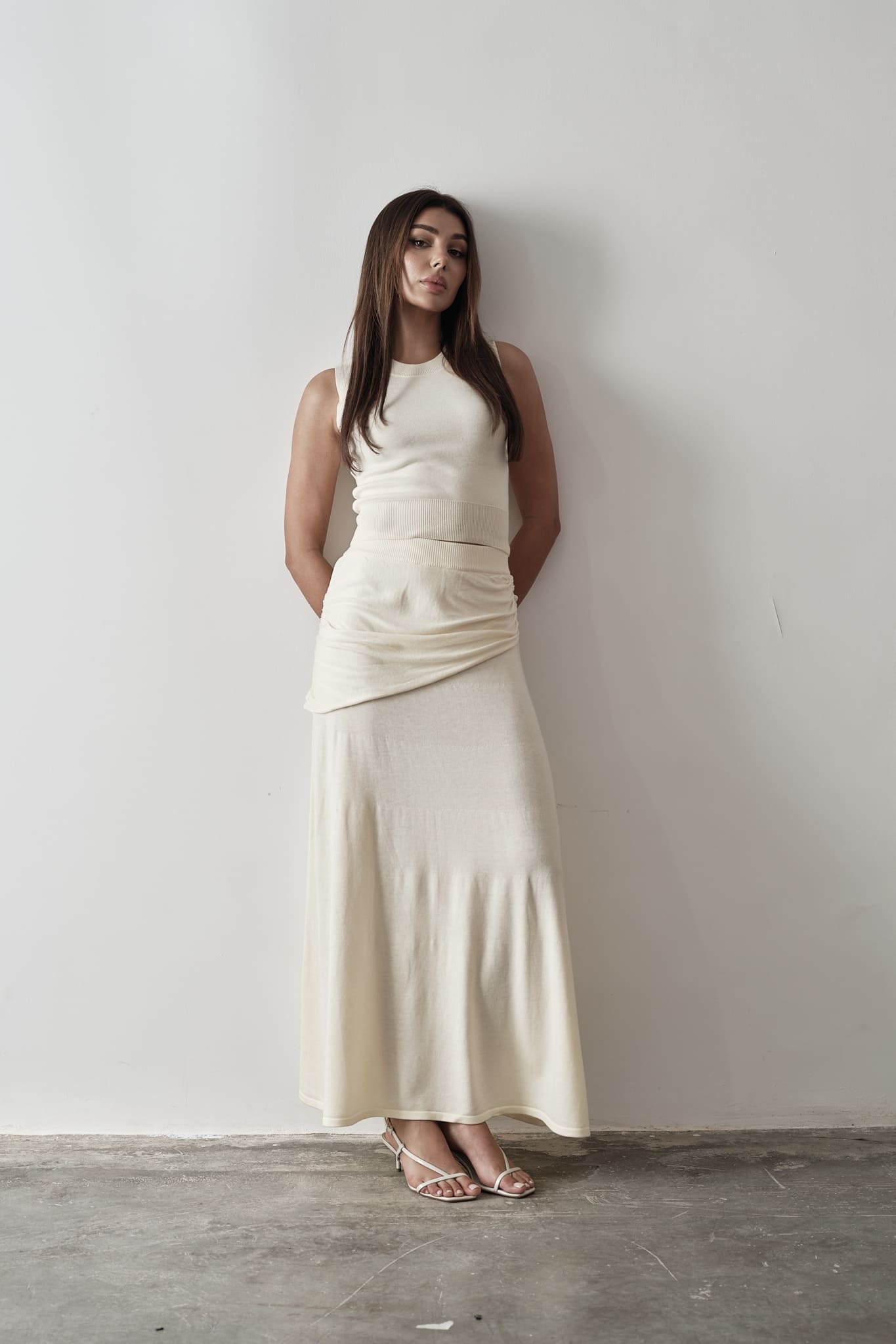 Model wearing a white Andrea Set featuring a sleeveless knit top with gathered detailing and a matching maxi skirt, styled with white strappy sandals, posing against a grey and white textured background