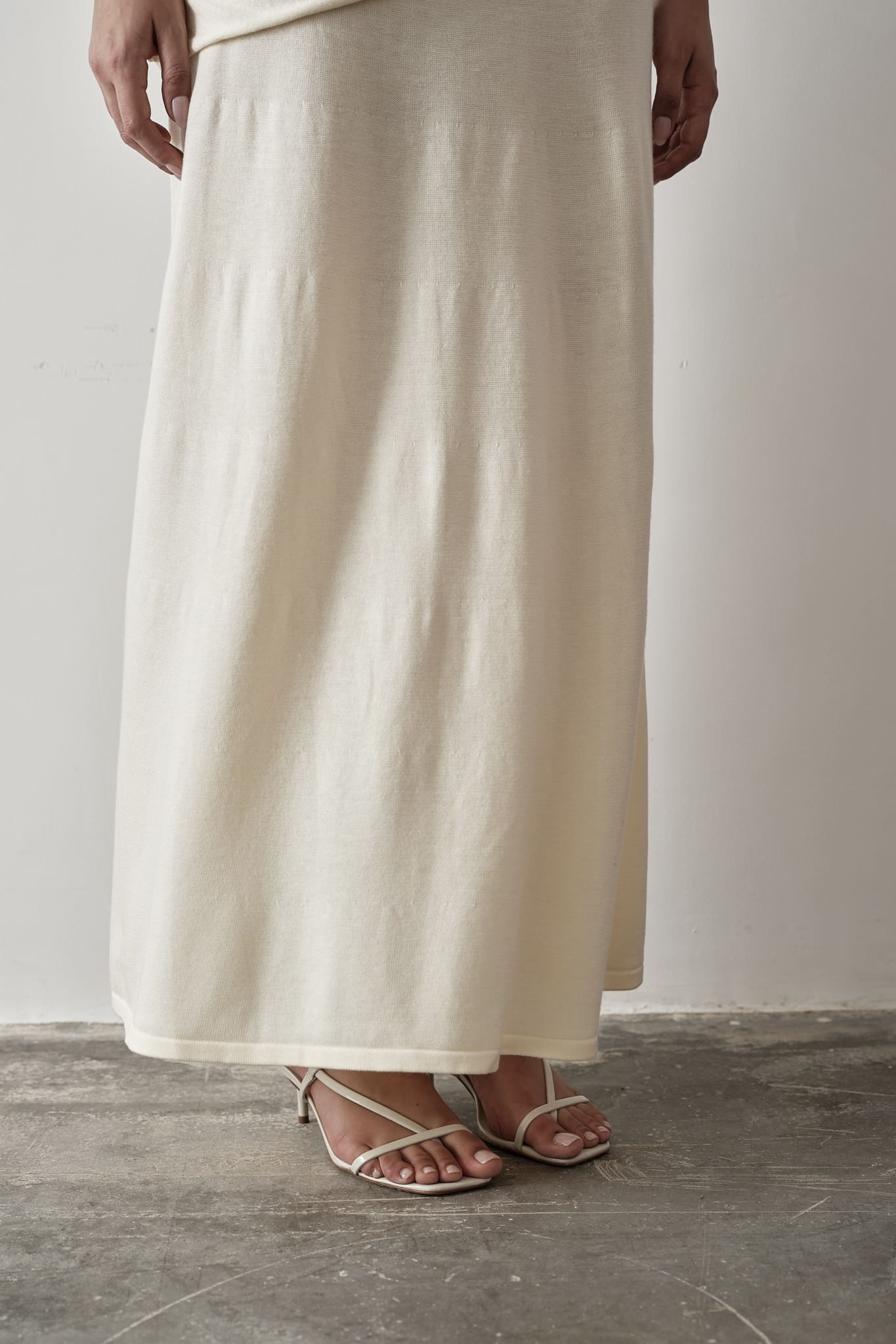 Model wearing a white Andrea Set featuring a sleeveless knit top with gathered detailing and a matching maxi skirt, styled with white strappy sandals, posing against a grey and white textured background