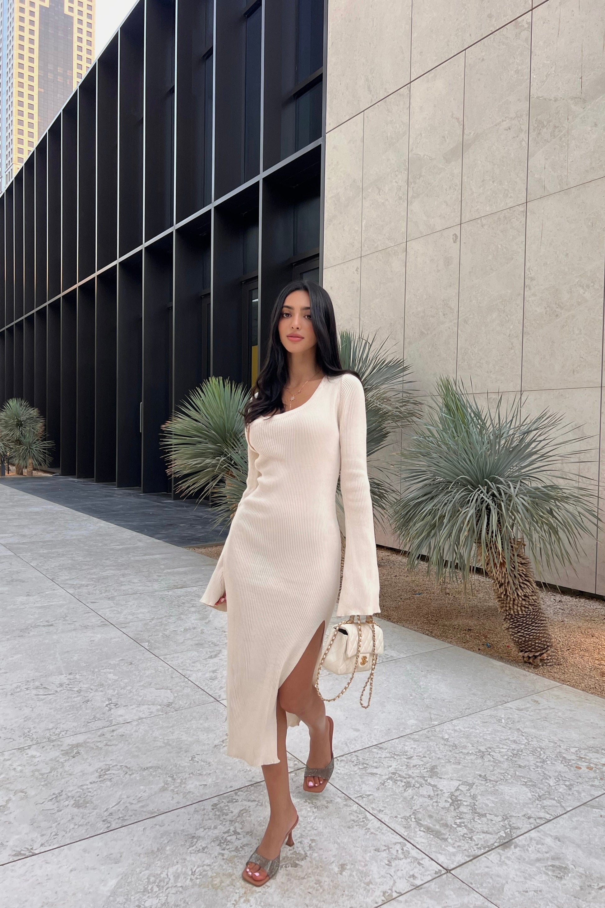 A woman in a cream Anja knit dress with a high slit stands outside a modern building, holding a small white handbag and wearing open-toe heels. Plants and a tiled walkway enhance the chic urban vibe.