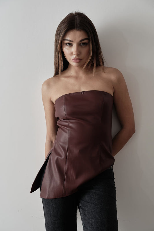 A woman stands against a plain background, modeling the Ellie leather top. The strapless design features a pleated dark green bodice paired with a sleek black skirt. Her dark hair is styled back, and she's completed the look with black heels, adopting a relaxed pose with one hand placed behind her back.