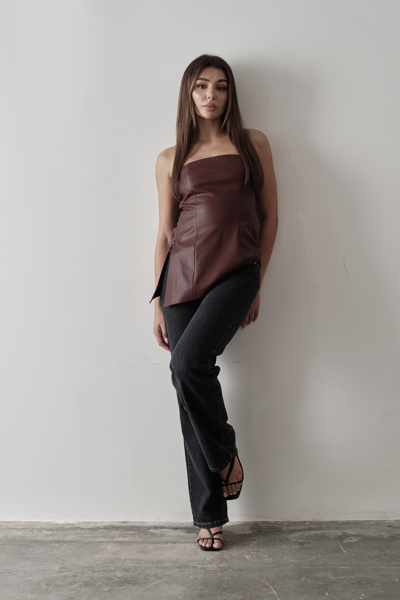 A woman stands against a plain background, modeling the Ellie leather top. The strapless design features a pleated dark green bodice paired with a sleek black skirt. Her dark hair is styled back, and she's completed the look with black heels, adopting a relaxed pose with one hand placed behind her back.