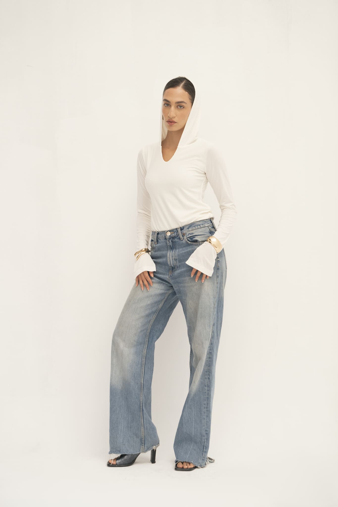 A person stands against a plain backdrop, dressed in a stylish Elyse Top featuring flared sleeves, paired with relaxed blue jeans. Their hands are casually tucked in the pockets of the jeans, and black open-toed heels add a touch of elegance, perfectly showcasing versatile functionality.