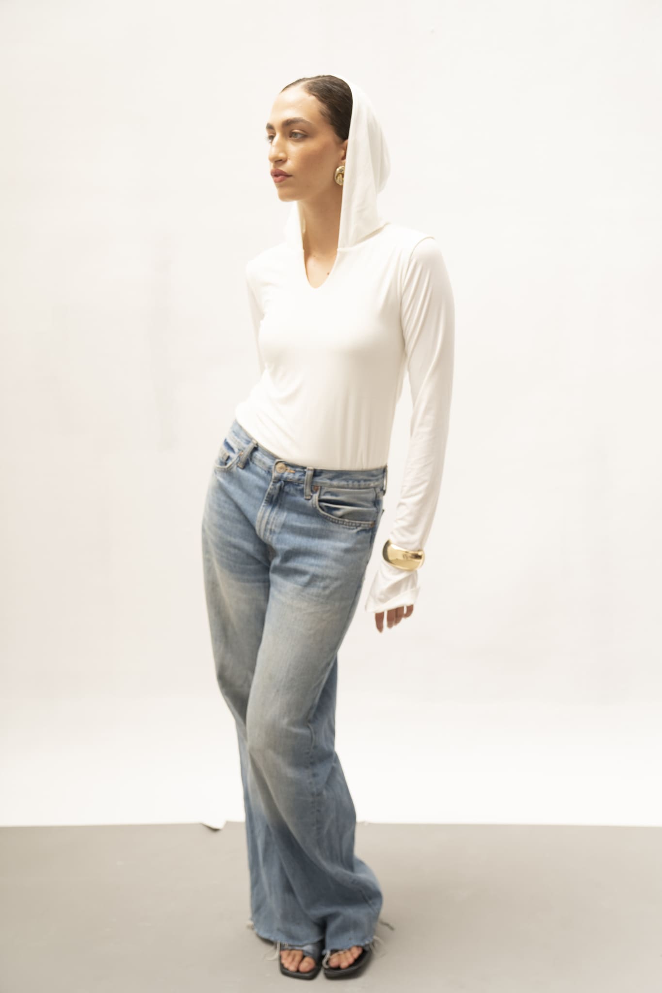 A person stands against a plain background wearing the sleek Elyse Top, blue jeans, and black footwear. With their head slightly turned to the side and one hand resting in their pocket, they exude versatile functionality. Their expression appears thoughtful.