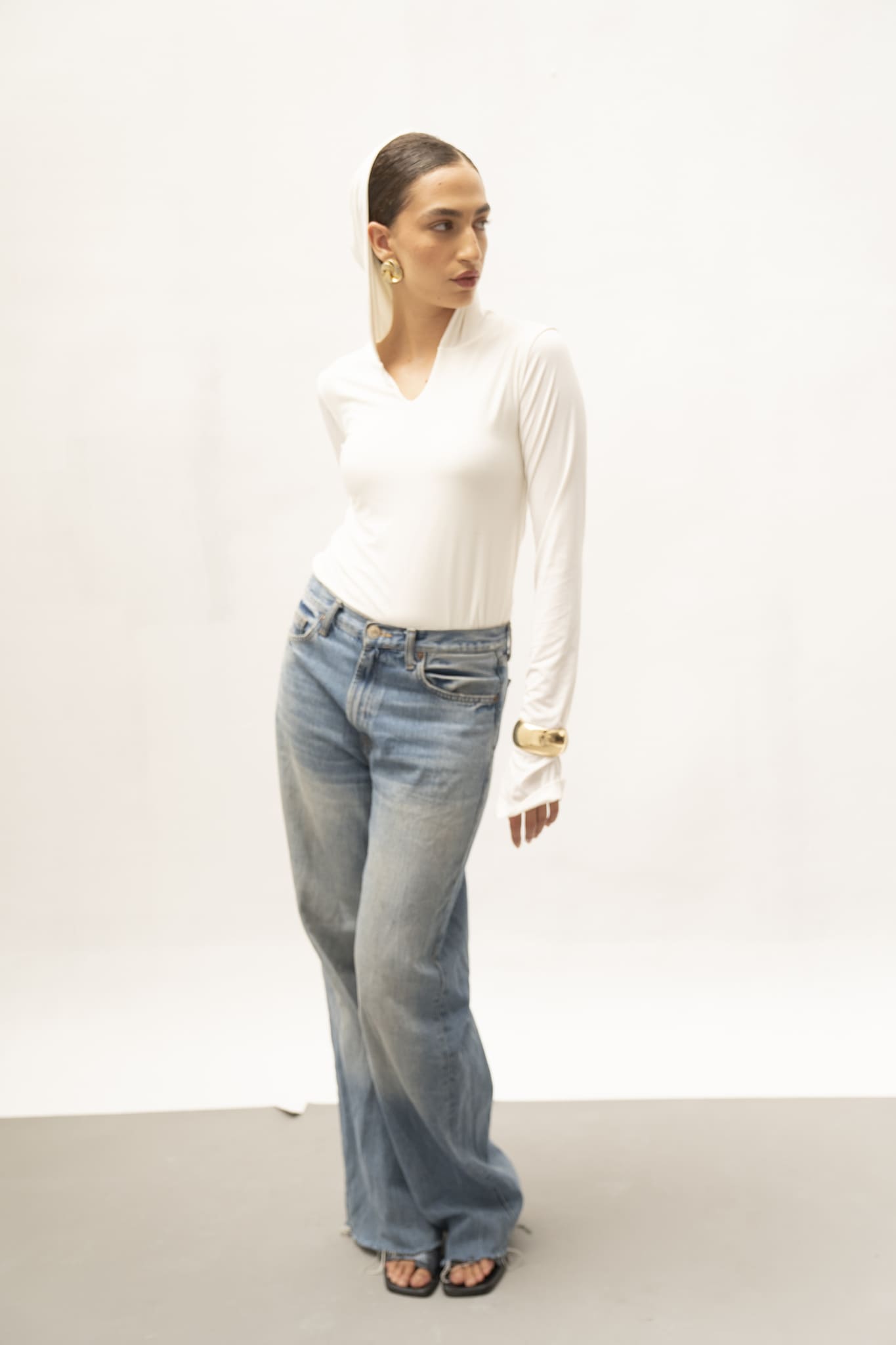 A person dressed in a white Elyse Top and blue jeans poses against a gray and white backdrop. They accessorize with large gold earrings and sandals, placing one hand on their hip while turning their head to the side, highlighting the Elyse Top's versatile functionality.