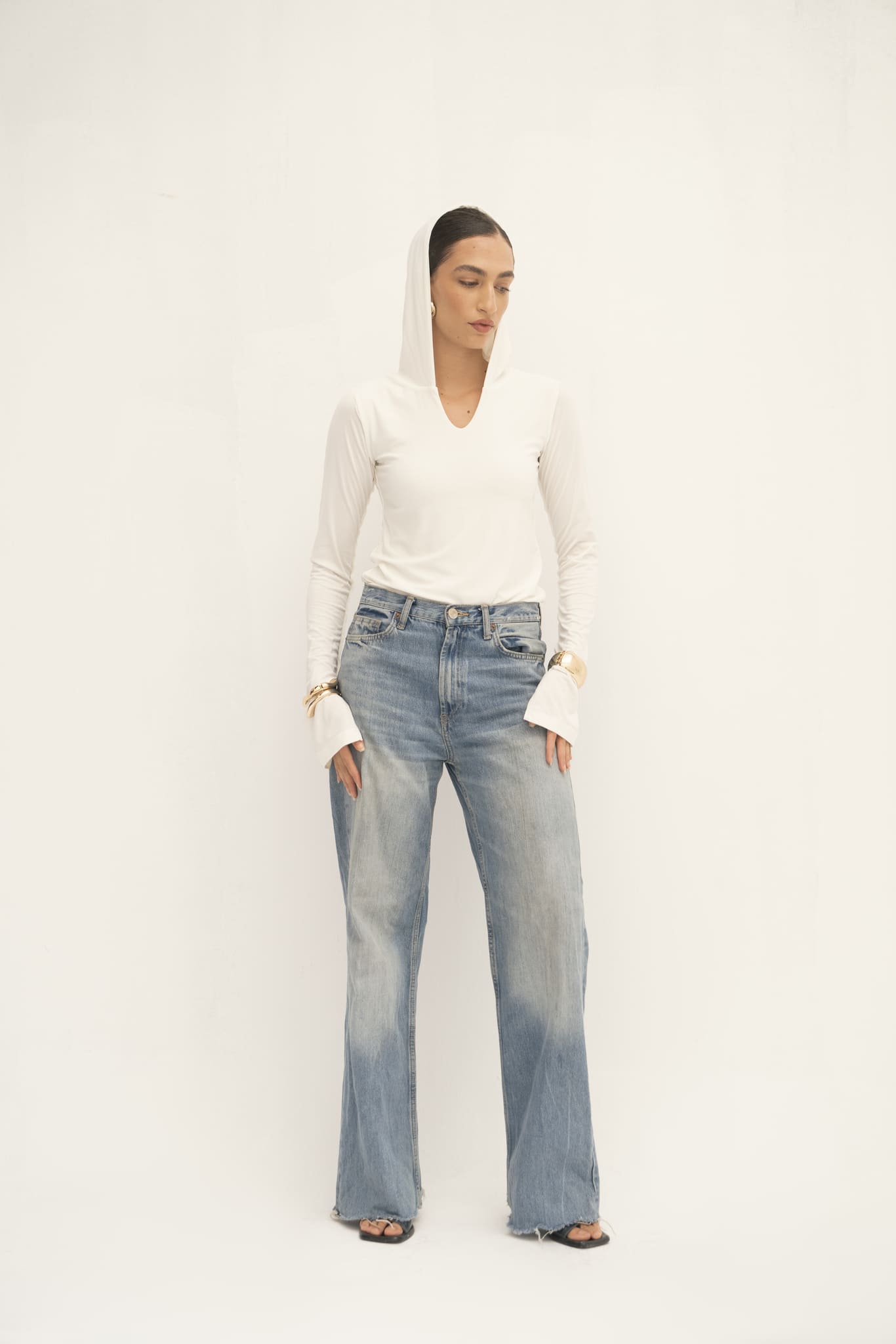 A person poses against a simple white backdrop, dressed in the stylish Elyse Top featuring a hood. They're paired with wide-leg jeans and sandals. Their long hair is pulled back, and they are adorned with gold bracelets.