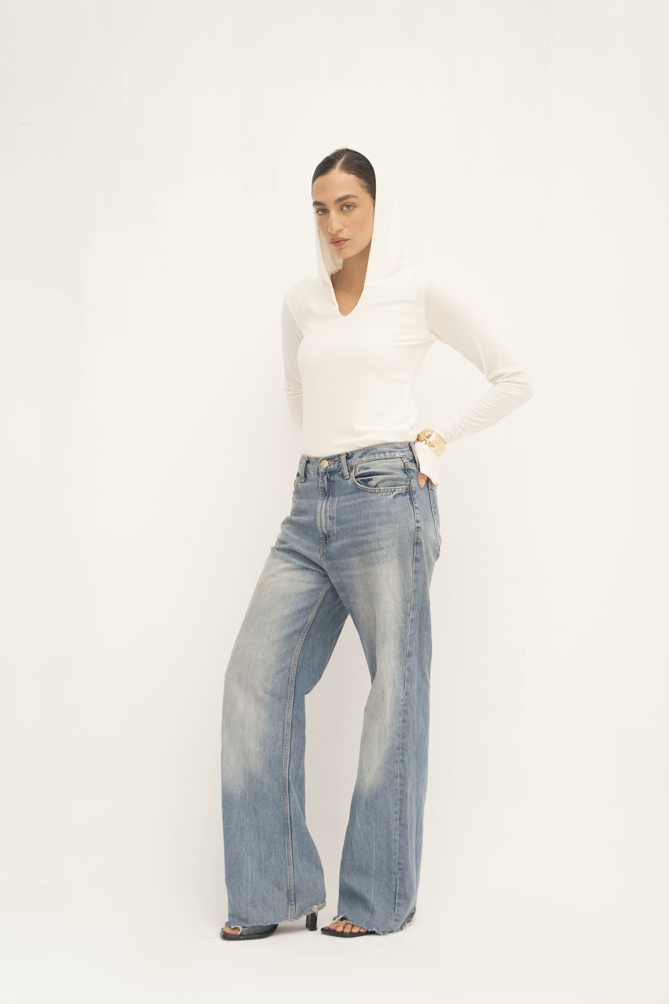 Dressed in the elegant Elyse Top paired with wide-leg blue jeans, the individual stands confidently against a light backdrop. They sport heels and rest a hand on their waist, emphasizing the stylish versatility of their chic outfit.