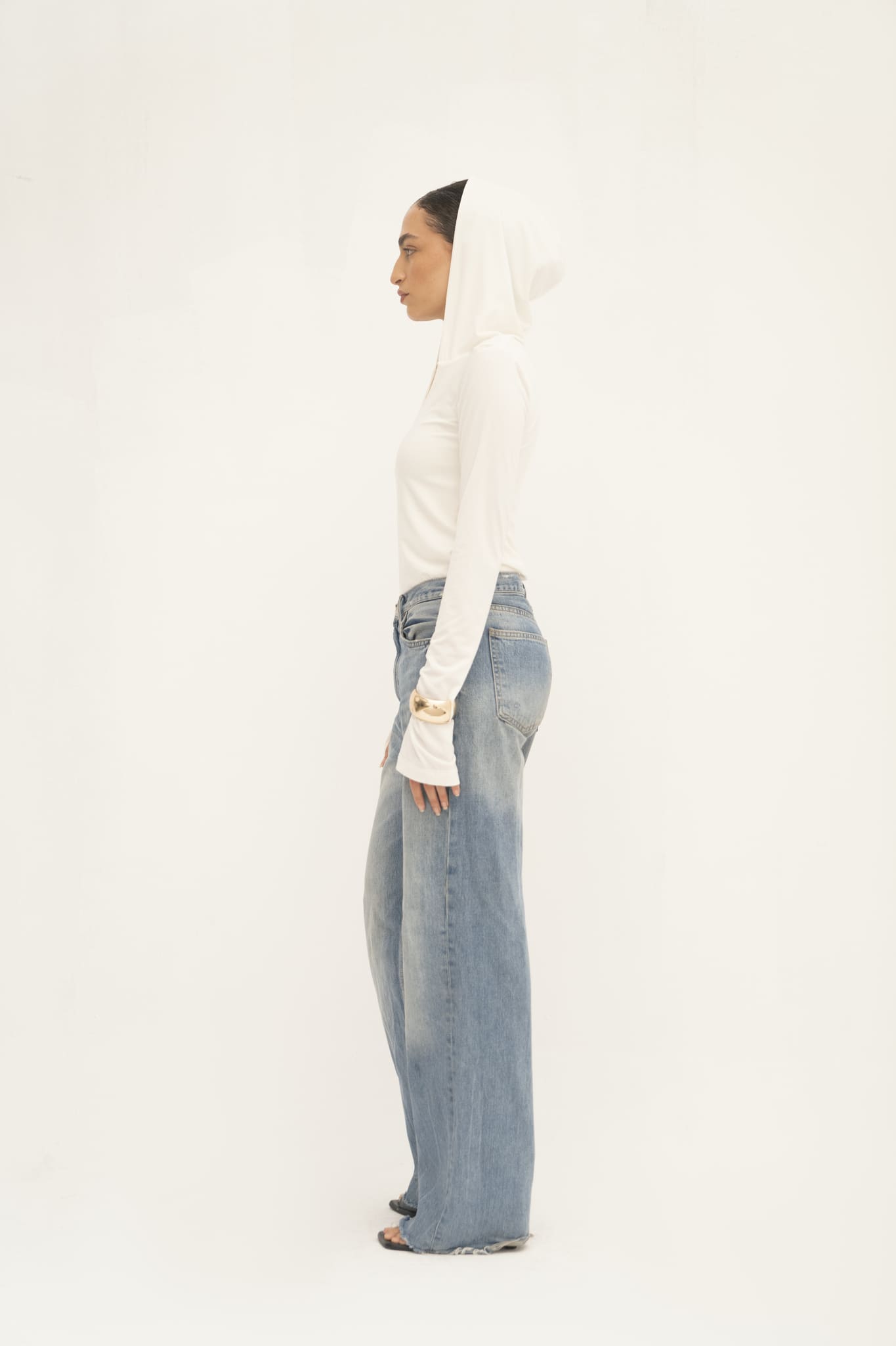 A person stands in profile against a plain white background, wearing the sleek Elyse Top and light blue wide-leg jeans. They appear relaxed, with their hair tucked inside the hood of the Elyse Top, showcasing its versatile functionality.