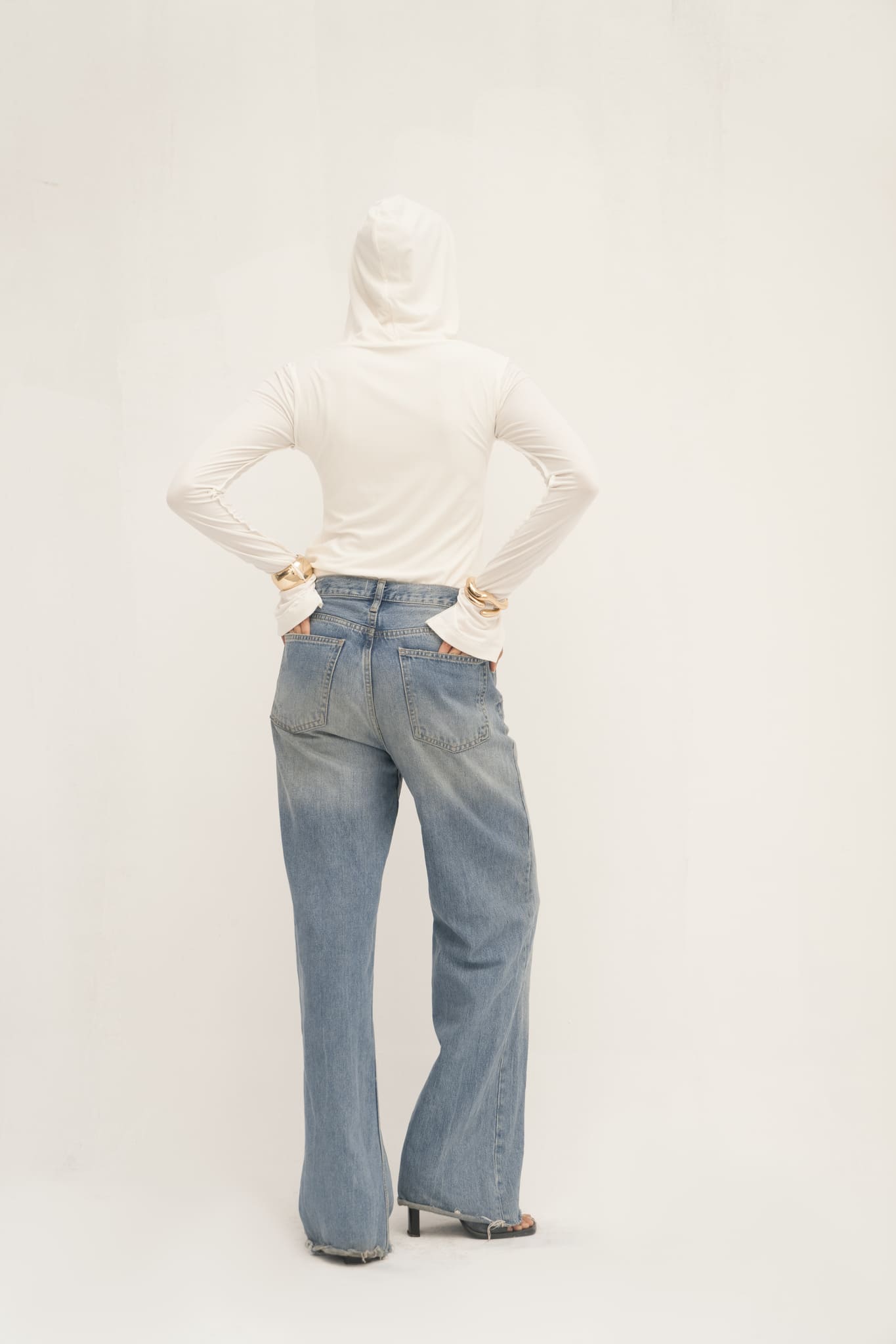 A person wearing the Elyse Top, a stylish white hoodie with a sleek hood, and blue jeans stands with their back to the camera. They have their hands on their hips and are wearing open-toed heels against a plain white background, highlighting versatile functionality in style.