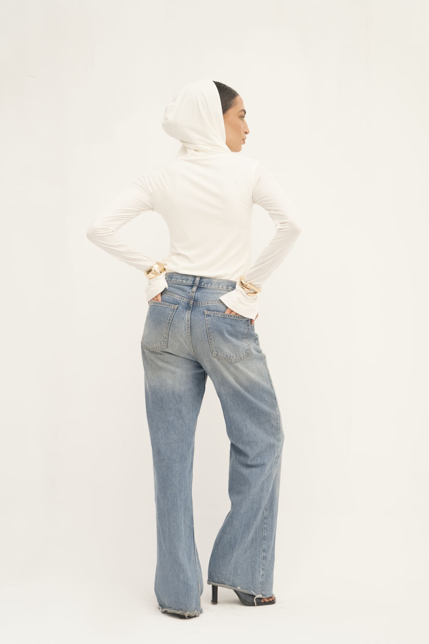 A person is seen facing away, highlighting the sleek hood of an Elyse Top in white, paired with blue wide-leg jeans. Their hands rest on their hips and they have dark hair. The minimalist white background emphasizes the outfit's versatile functionality.