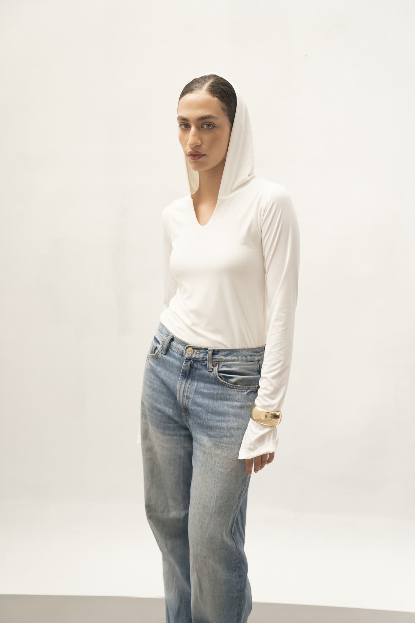 An individual stands in front of a simple backdrop dressed in the Elyse Top, a white long-sleeve hoodie noted for its sleek design, paired with blue jeans. Their hands rest in their pockets, complemented by a gold bracelet.