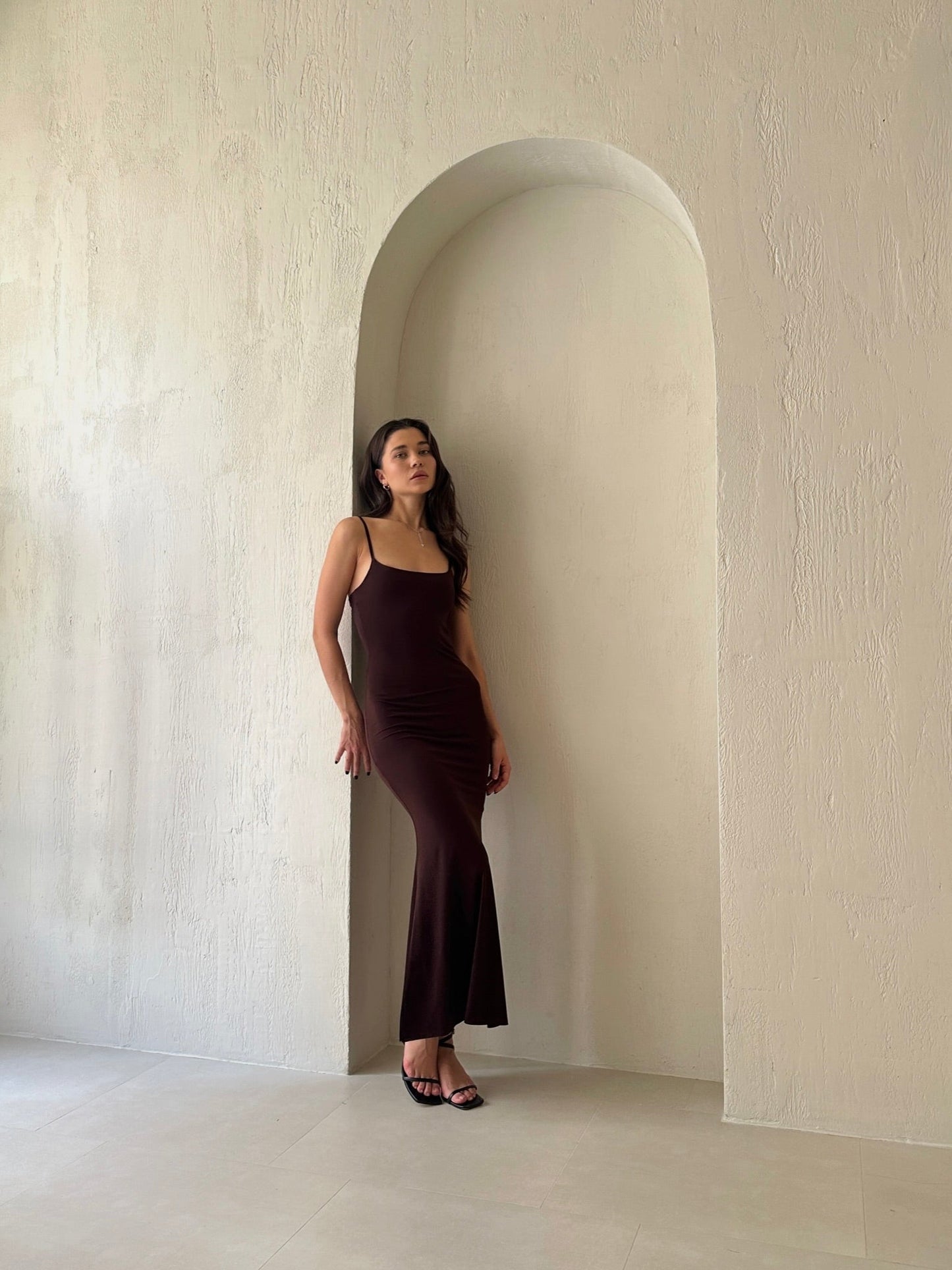 A woman in the Etta dress, a long black slip dress featuring adjustable spaghetti straps, stands against a smooth, neutral-toned arched wall. She gazes upward with her hair down, embodying a minimalist and elegant aesthetic.