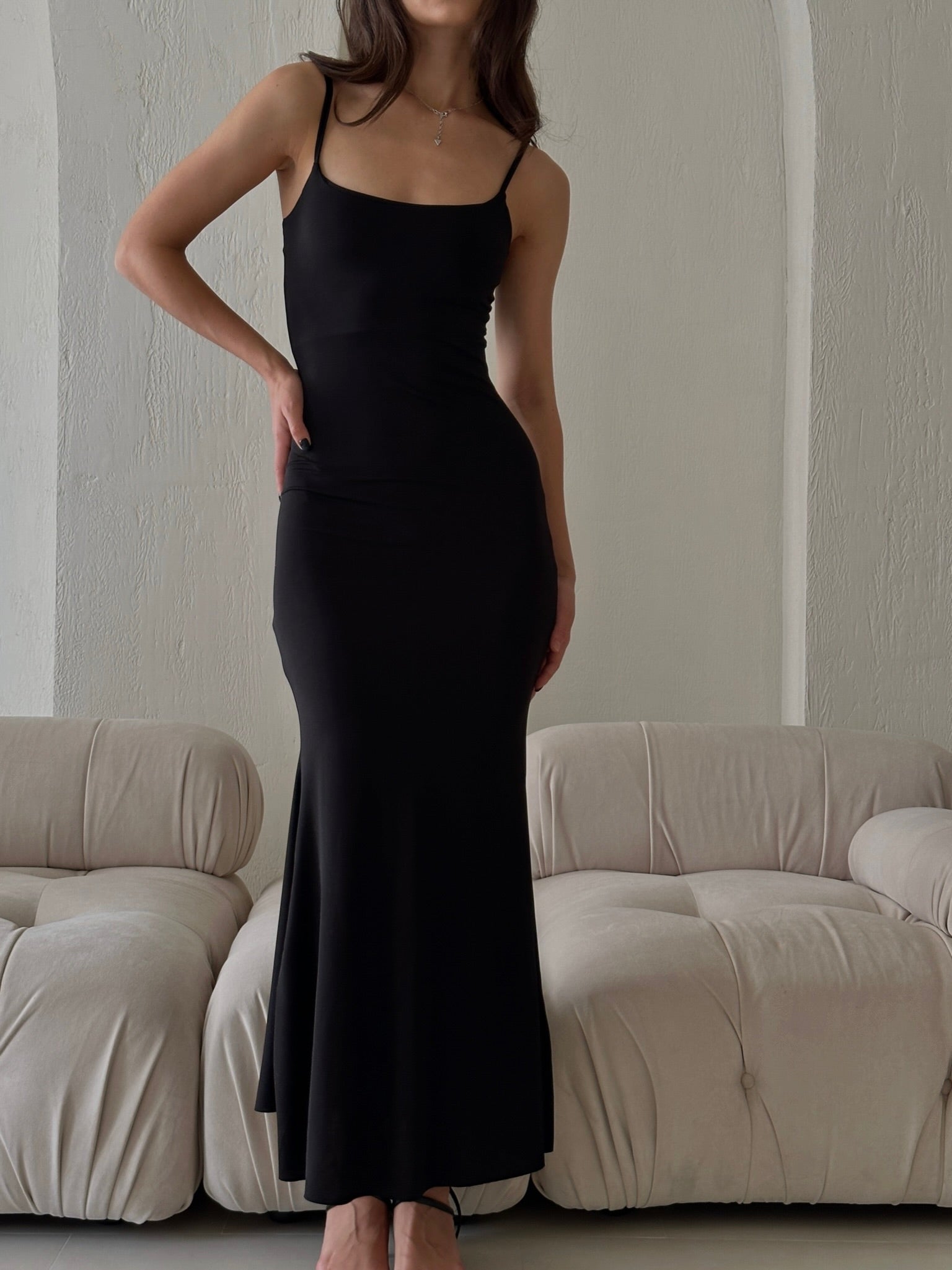 Wearing the Etta dress, a person stands in a room featuring a cream-colored couch. The long, fitted black slip dress with adjustable spaghetti straps beautifully accentuates their figure. Their face remains unseen against the backdrop of a textured, light-colored wall.