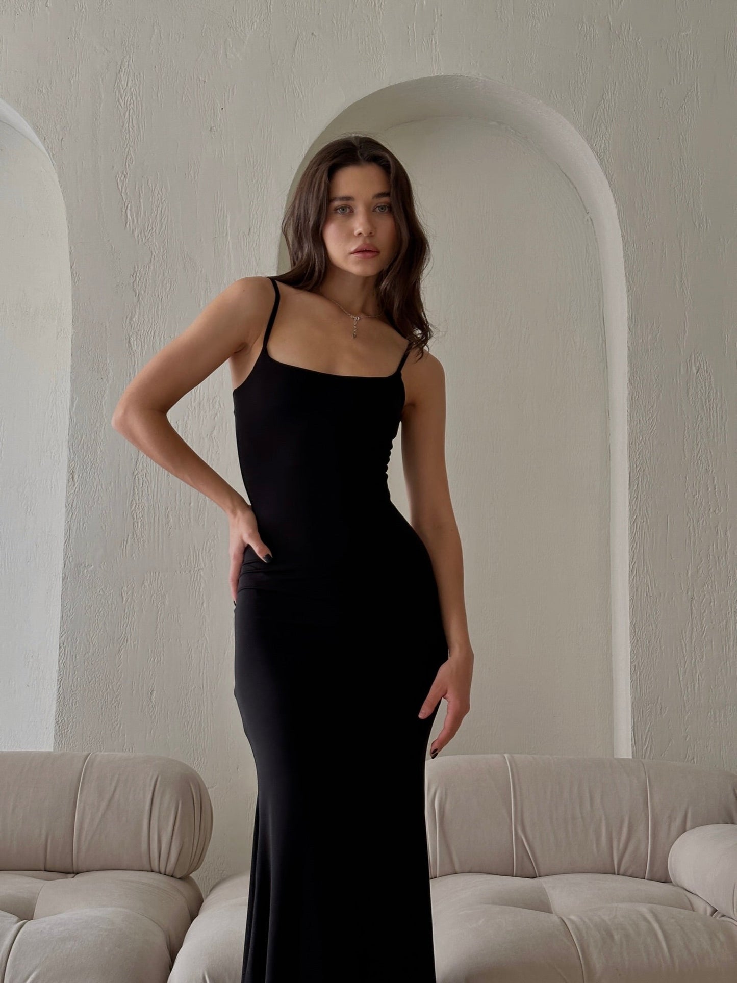 A woman in the elegant Etta dress, featuring sleek adjustable spaghetti straps, poses gracefully in front of a textured light-colored wall with arched cutouts. She stands between beige tufted sofas, exuding an air of sophistication. Experience international delivery for this stunning look.