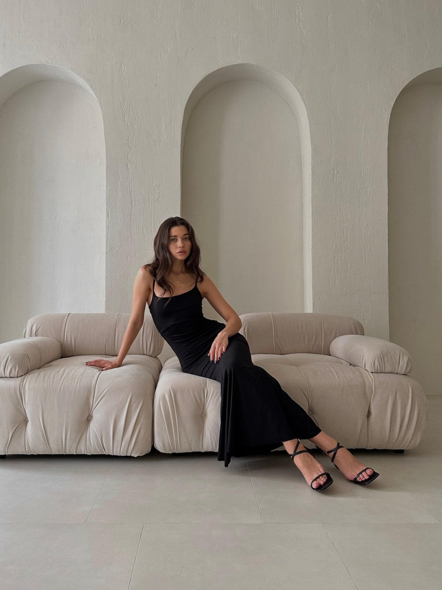 A woman wearing the Etta dress, a long black slip, sits on a beige tufted sofa placed against a minimalist white wall with three arches. Her long, dark hair cascades down as she sports strappy black heeled sandals.