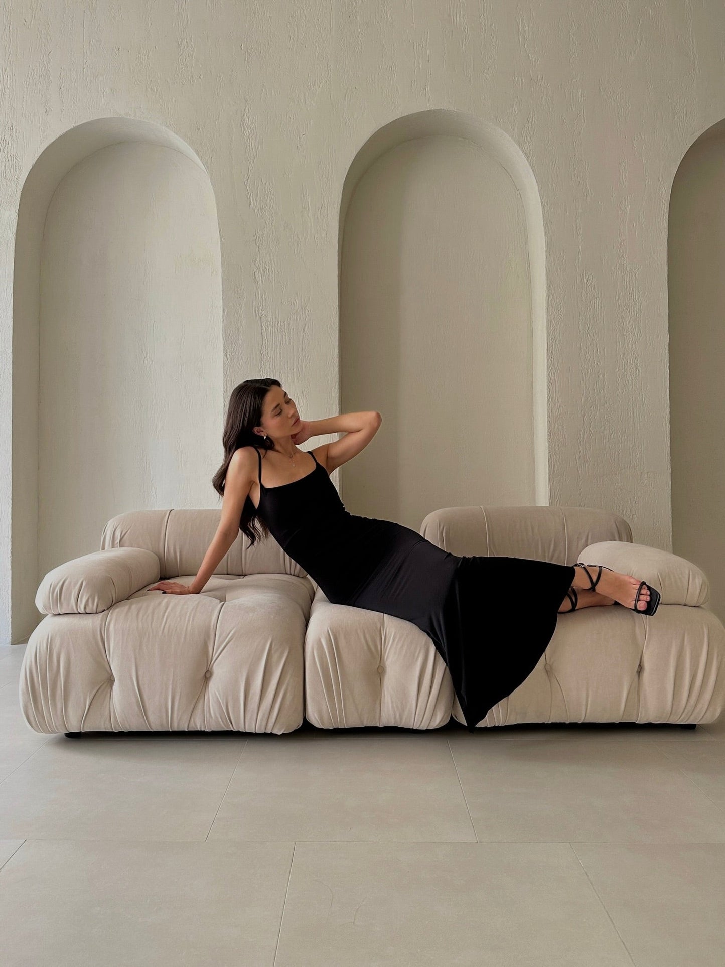 A woman wearing the stylish Etta dress is elegantly reclining on a plush, cream-colored sofa. The background features textured, arched alcoves that add depth to the minimalist setting crafted from high-quality materials.