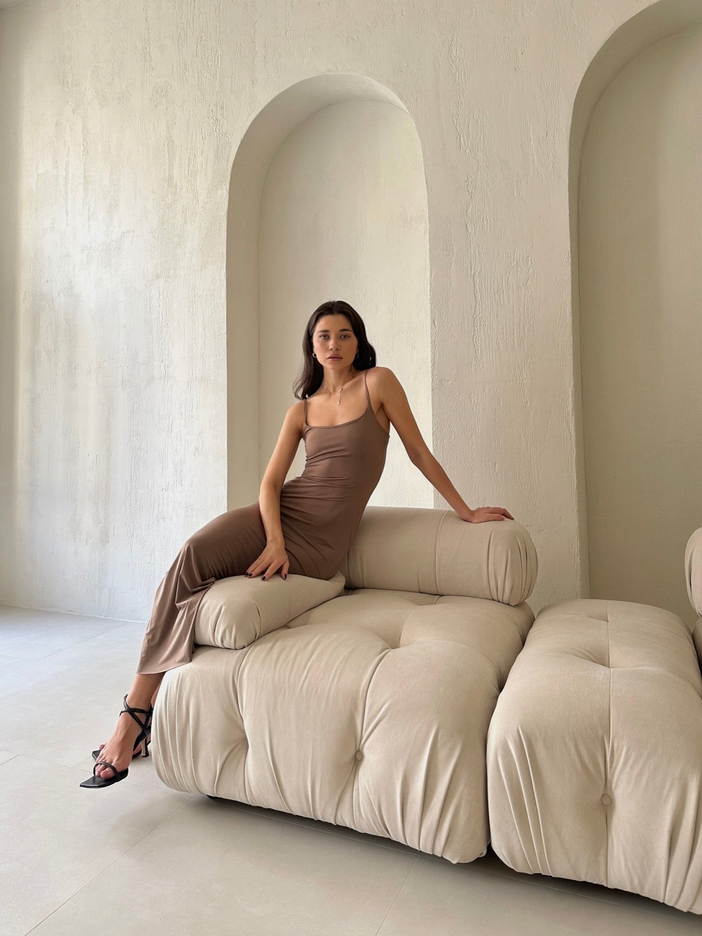 A woman in the elegant Etta dress, a sleek brown slip-style number, is seated on a modern cream-colored sofa set against a textured wall with arched niches. She rests one hand on her knee and the other on the back of the sofa. Wearing black heels and gazing directly at the camera, she emphasizes the high-quality materials of her attire.