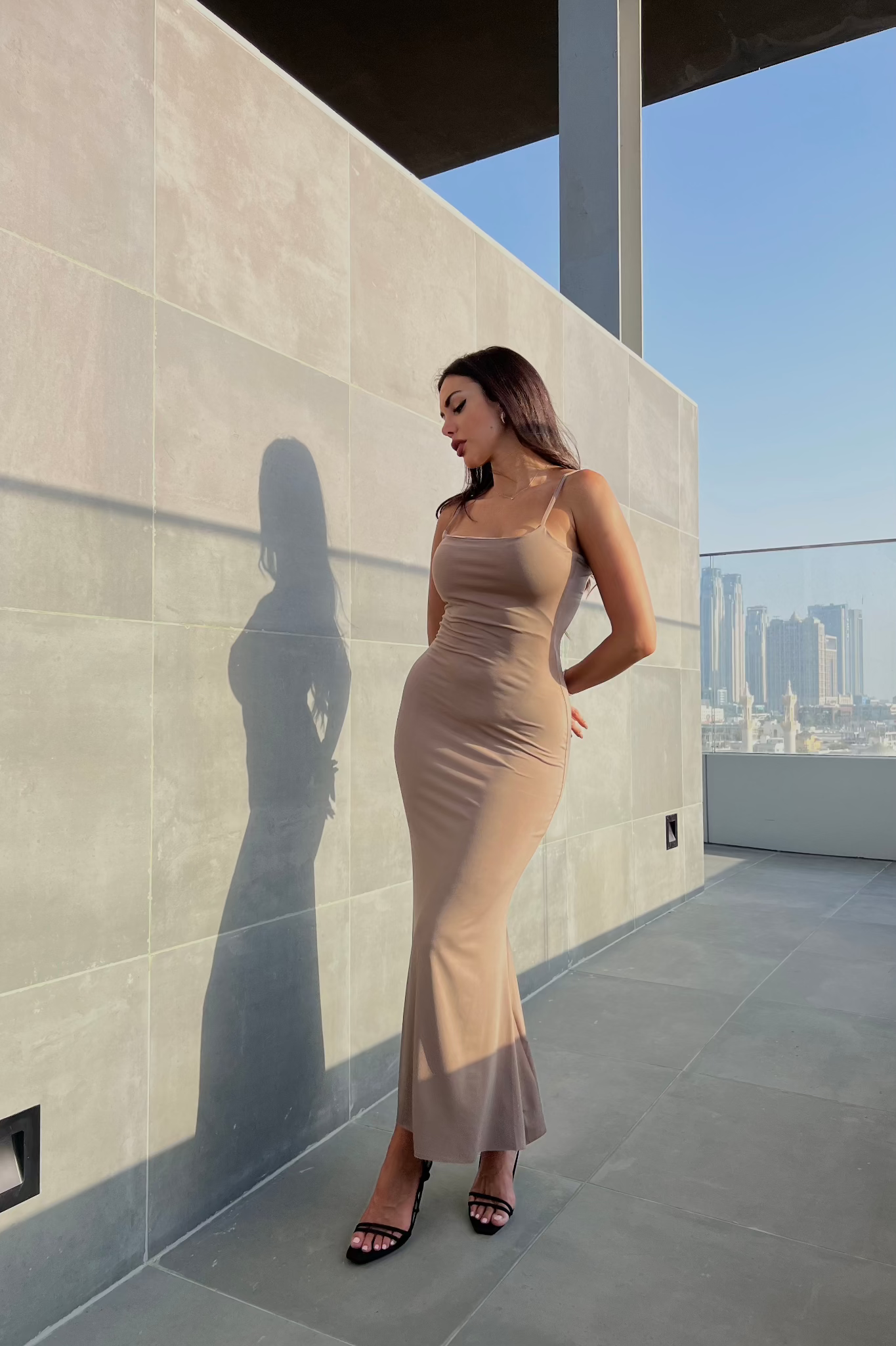 Dressed in the Etta dress, a woman stands on a balcony with a city skyline behind her. The fitted slip dress, made from high-quality materials, highlights her silhouette as she casts a shadow on the wall. She completes her modern and urban look with black heeled sandals.