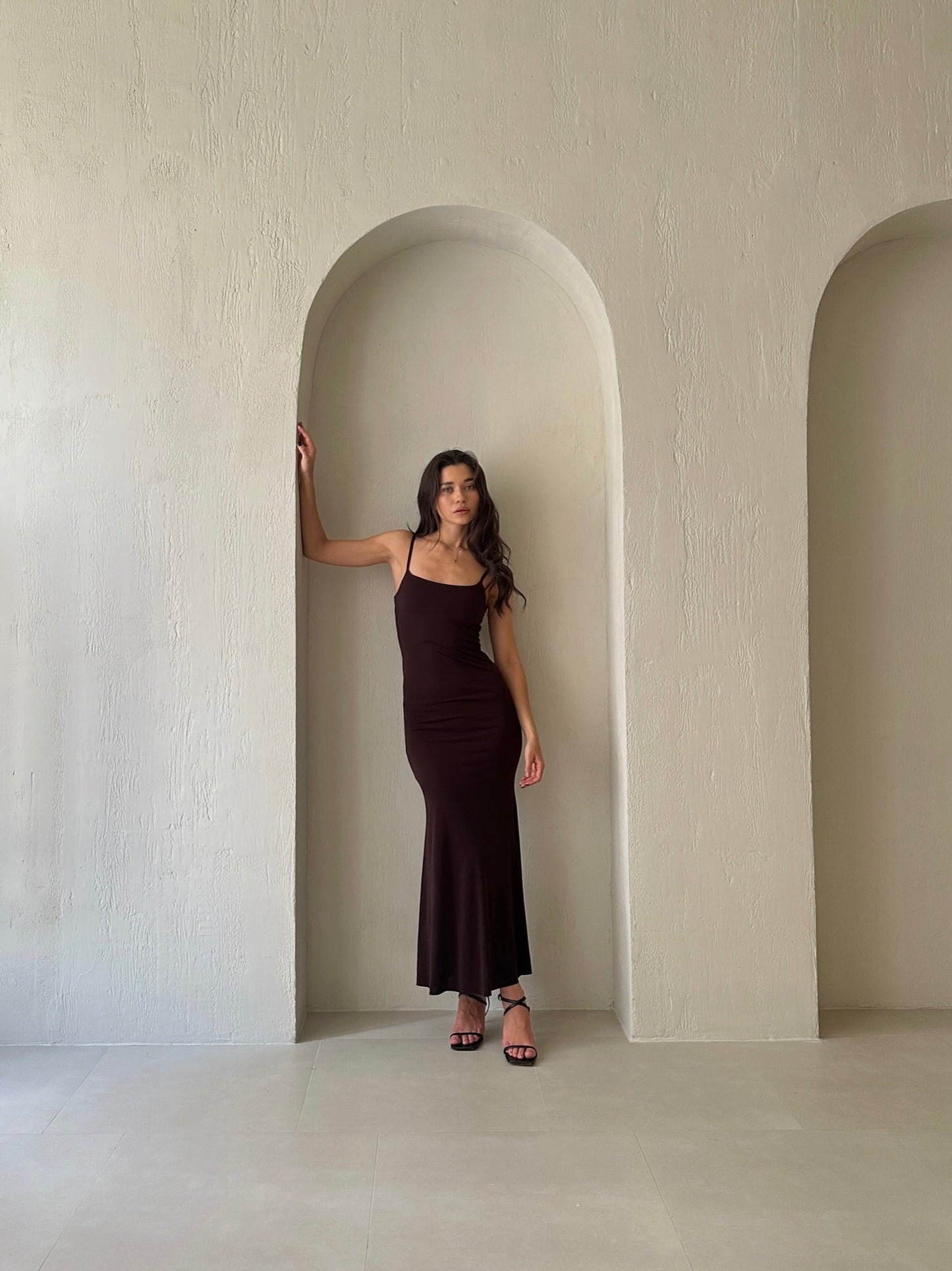 A person wearing the Etta dress, which features a long, dark slip design and adjustable spaghetti straps, stands in a minimalist arch-shaped alcove, paired with black strappy heels. The plain, textured wall enhances the simplicity and elegance of the setting.