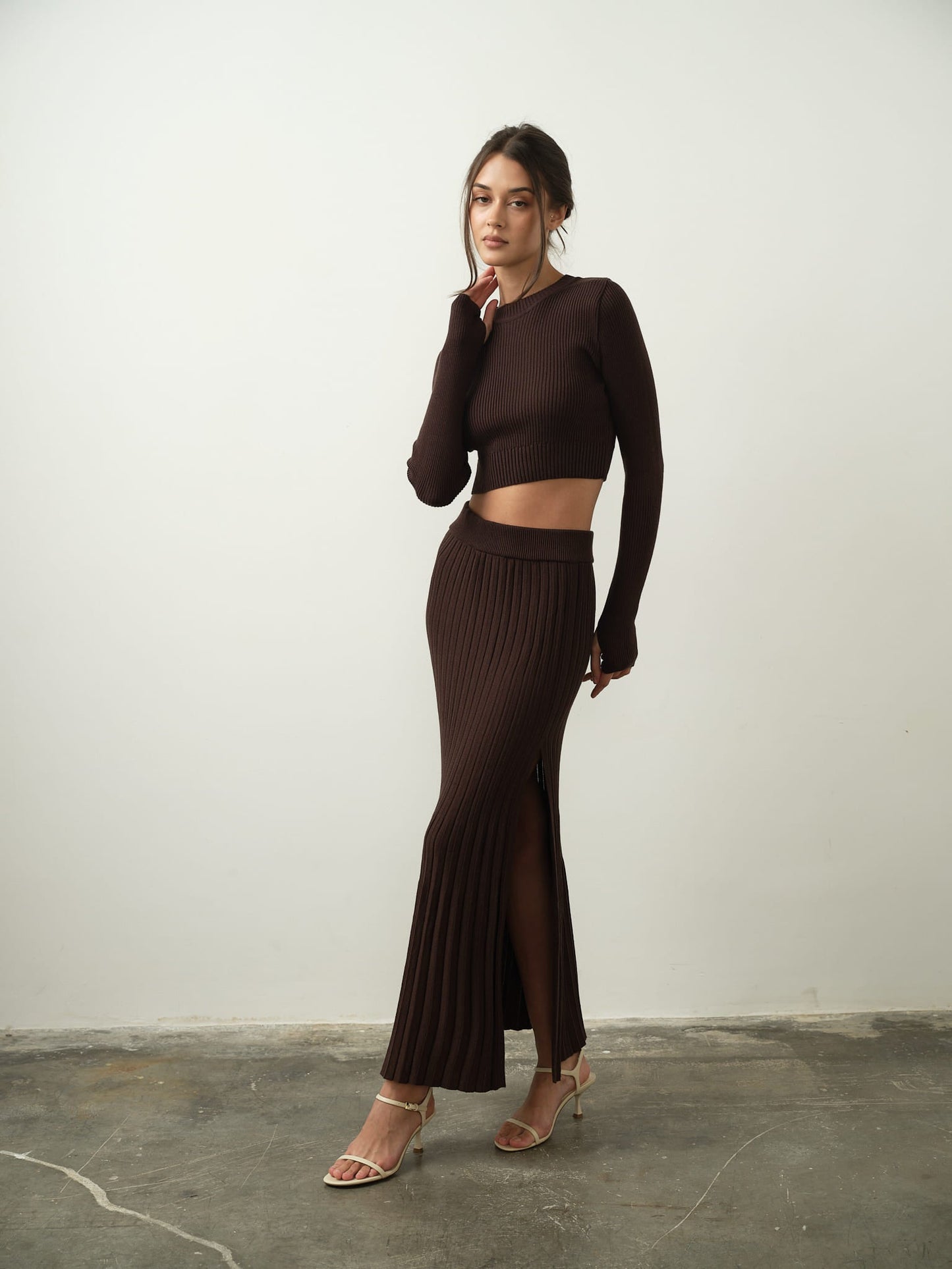A person poses against a plain background, wearing the Gaïa Set—a brown long-sleeve crop top paired with a matching high-waisted maxi skirt featuring a slit—crafted from high-quality cotton knit fabric. They stand on a concrete floor, gazing at the camera with one hand touching their face, exuding modern femininity.