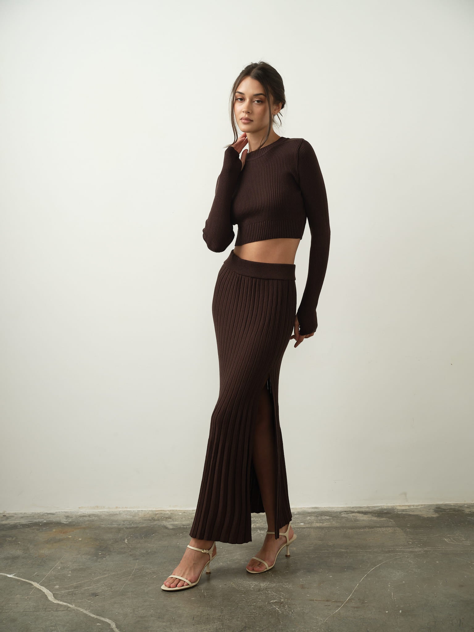 A person poses against a plain background, wearing the Gaïa Set—a brown long-sleeve crop top paired with a matching high-waisted maxi skirt featuring a slit—crafted from high-quality cotton knit fabric. They stand on a concrete floor, gazing at the camera with one hand touching their face, exuding modern femininity.