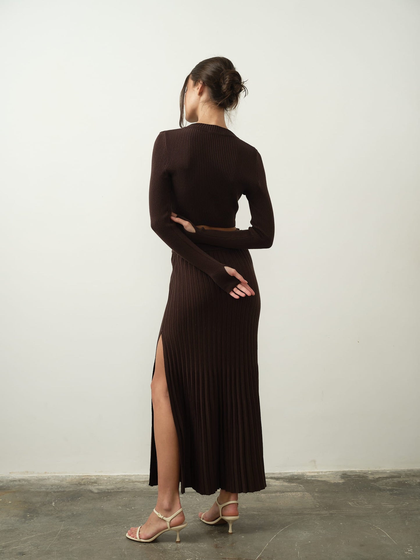A person stands with their back to the camera, wearing the Gaïa Set—a long, dark ribbed dress crafted from high-quality cotton knit fabric. Their hair is styled up, and they hold one arm behind them. The dress features a slit, complemented by strappy heels, capturing modern femininity against a plain background.