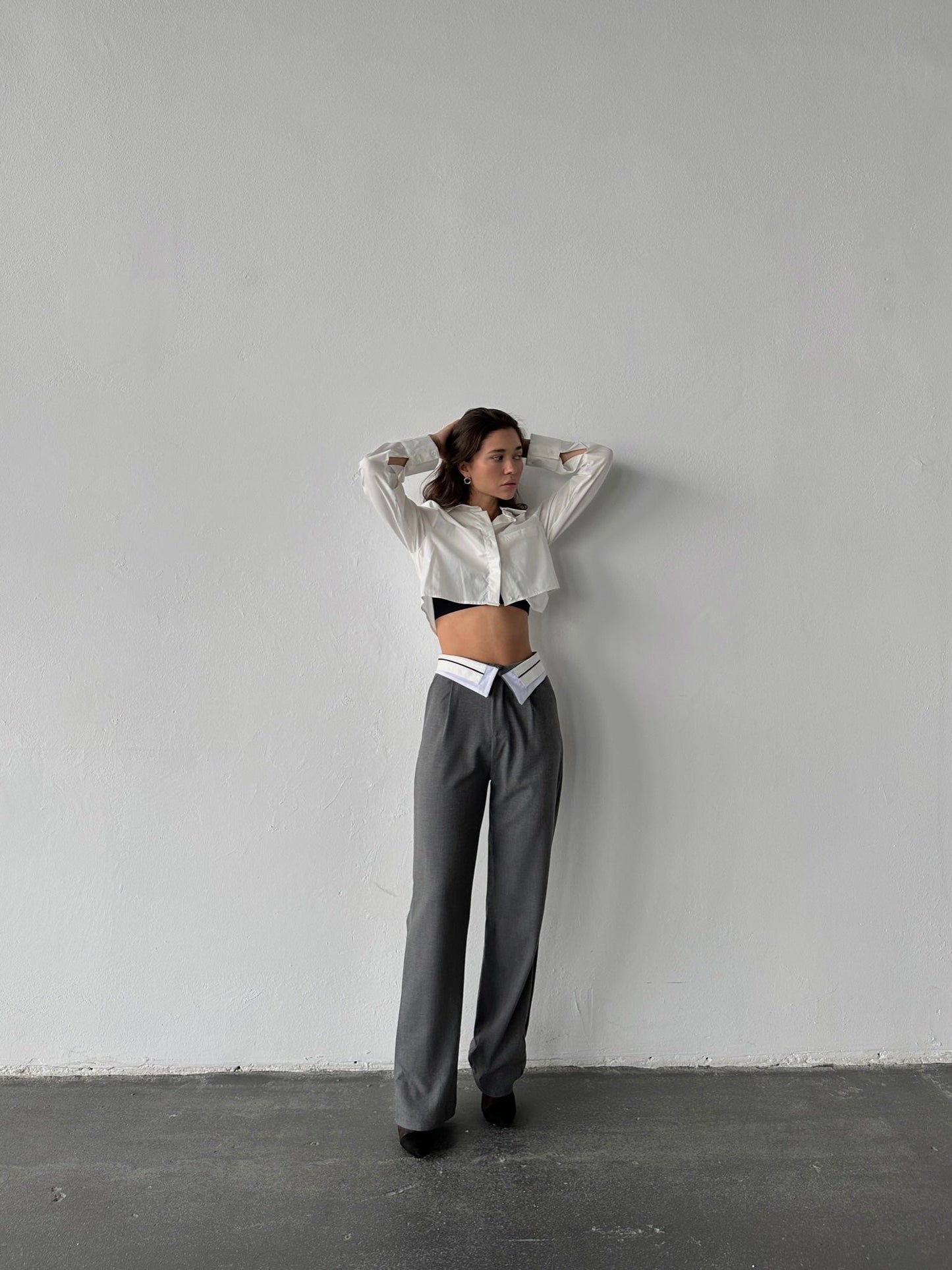 Standing against a plain wall, they wear a white cropped blouse and Rina Pants—gray wide-leg trousers with a unique waistband cut. With arms raised and hands on their head, they gaze to the side. The breathable fabric enhances the comfort of this chic ensemble.
.