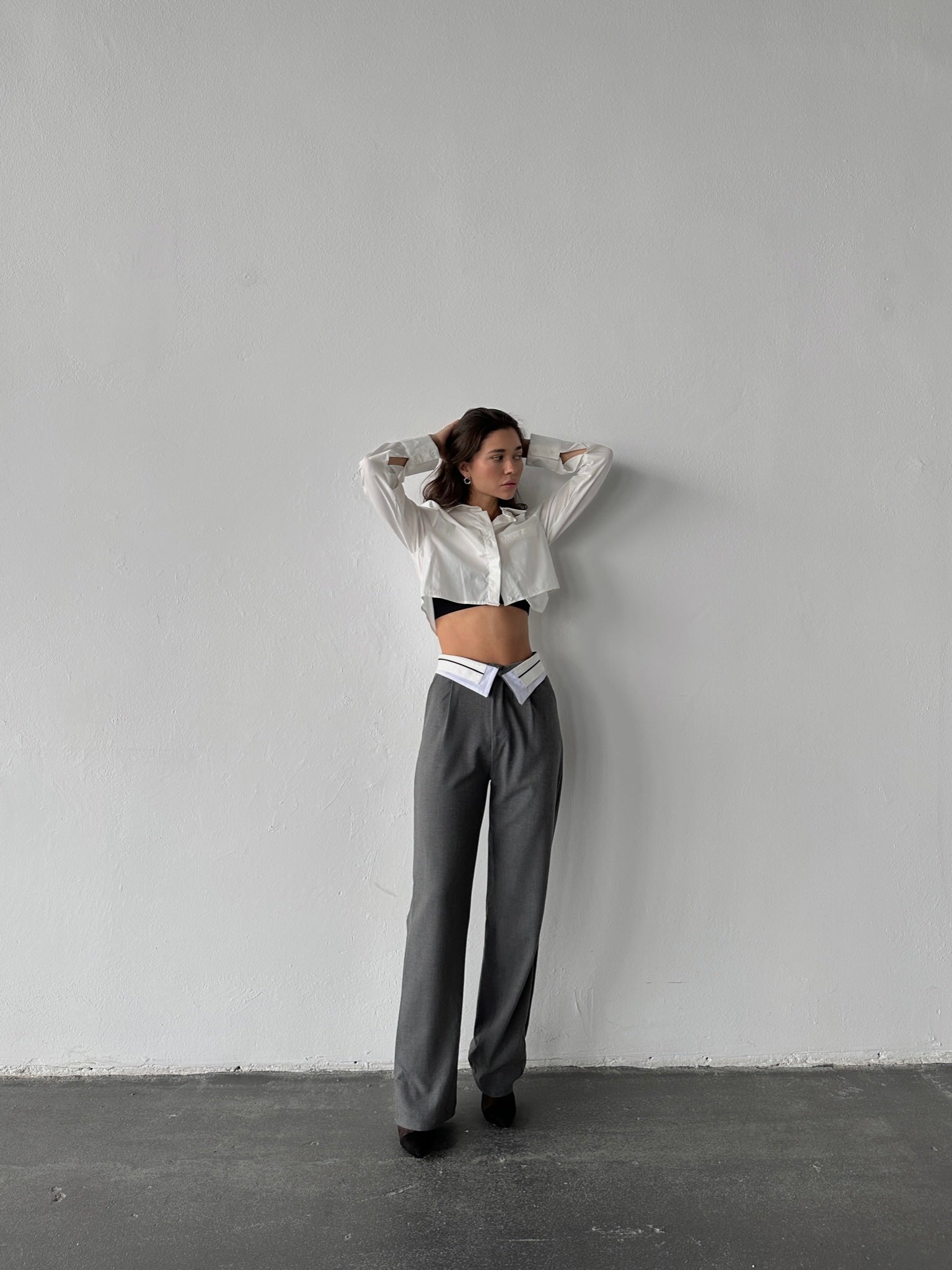 Standing against a plain wall, they wear a white cropped blouse and Rina Pants—gray wide-leg trousers with a unique waistband cut. With arms raised and hands on their head, they gaze to the side. The breathable fabric enhances the comfort of this chic ensemble.
.