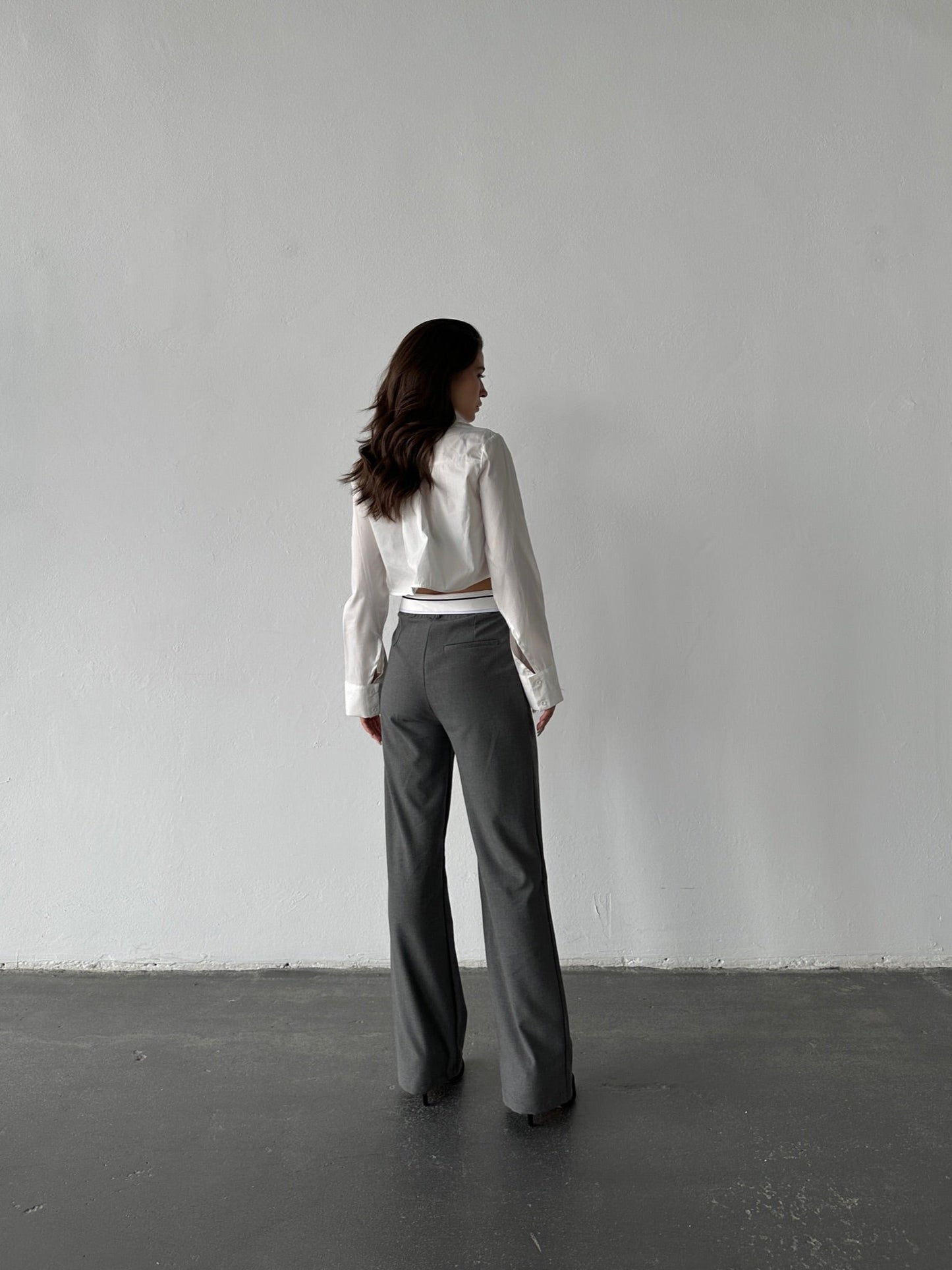 A person with long dark hair stands against a plain, light-colored wall, wearing a white long-sleeve shirt and gray Rina Pants in a breathable fabric. They look away from the camera as the dark-colored floor contrasts with the wall.