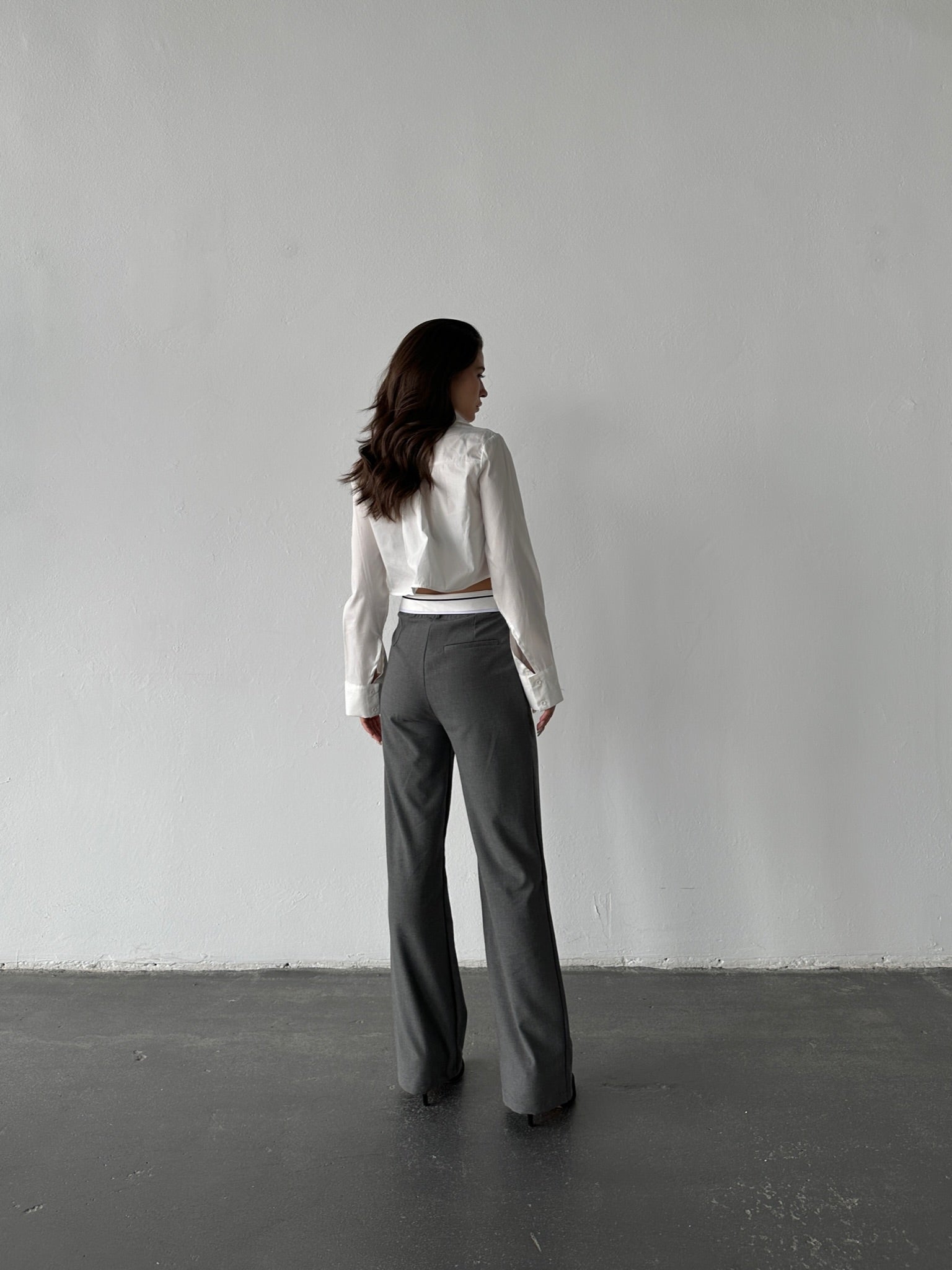 A person with long dark hair stands against a plain, light-colored wall, wearing a white long-sleeve shirt and gray Rina Pants in a breathable fabric. They look away from the camera as the dark-colored floor contrasts with the wall.