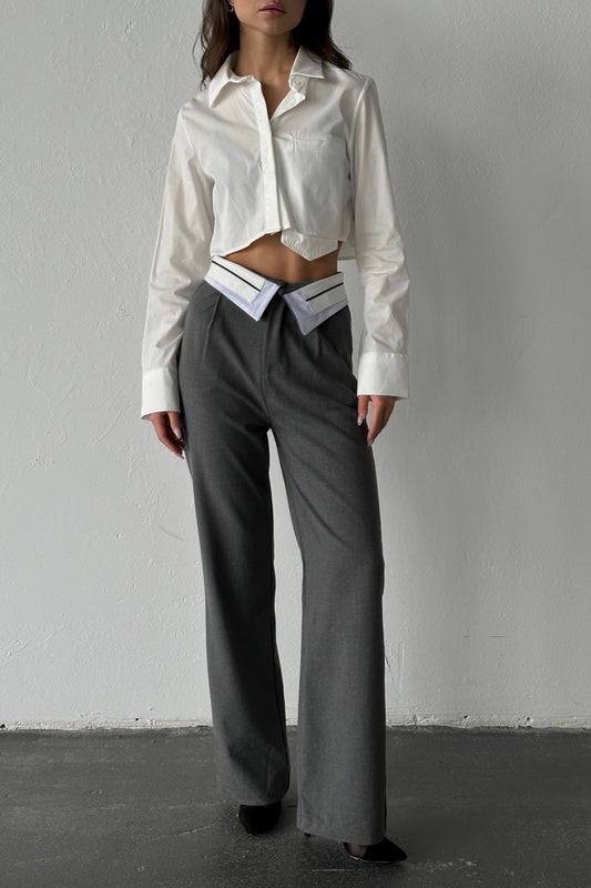 A person stands against a white wall, wearing a cropped white button-up shirt and high-waisted Rina Pants in gray. The breathable fabric of the wide-leg pants is highlighted as they rest their hands at their sides on the dark floor.