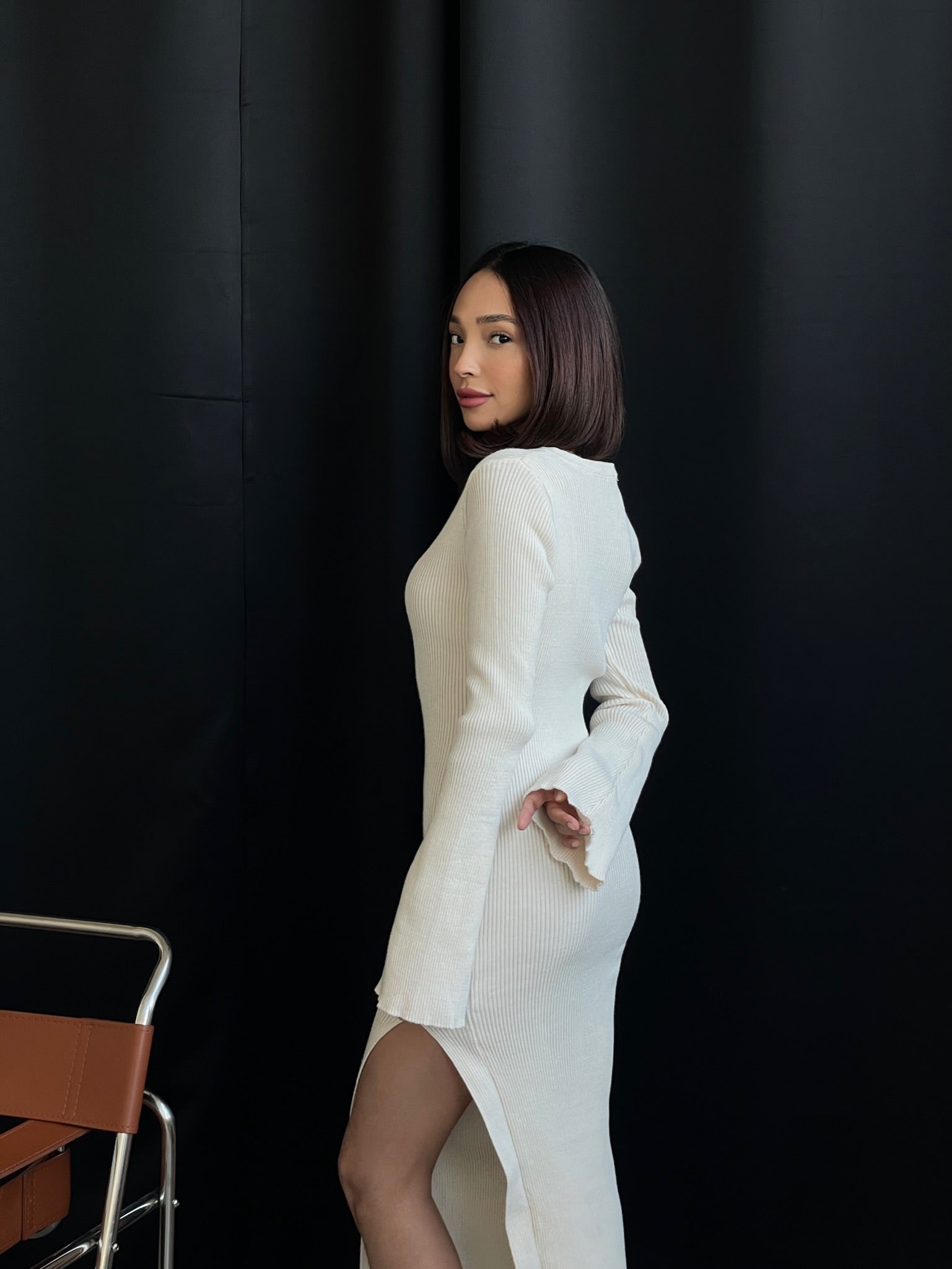 A person with shoulder-length dark hair stands sideways, turning slightly towards the camera. They wear the Anja knit dress, a long-sleeved white midi with a high slit. A black curtain is in the backdrop, and part of a brown chair appears to the left.