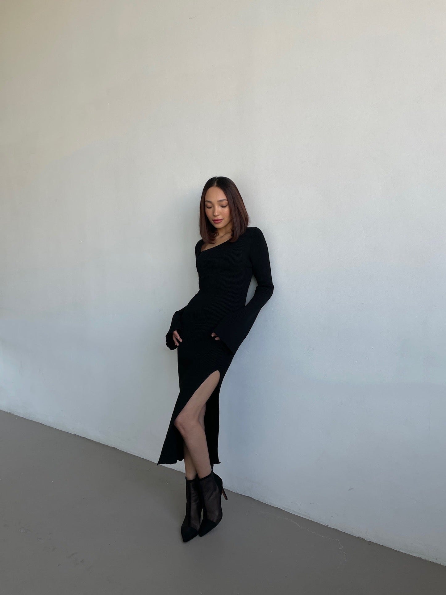 A woman wearing the Anja knit dress, a long-sleeved black midi with a high slit, stands against a plain wall. Her straight hair rests on her shoulders, and she's in black ankle boots. She poses with one hand on her hip and the other at her side.