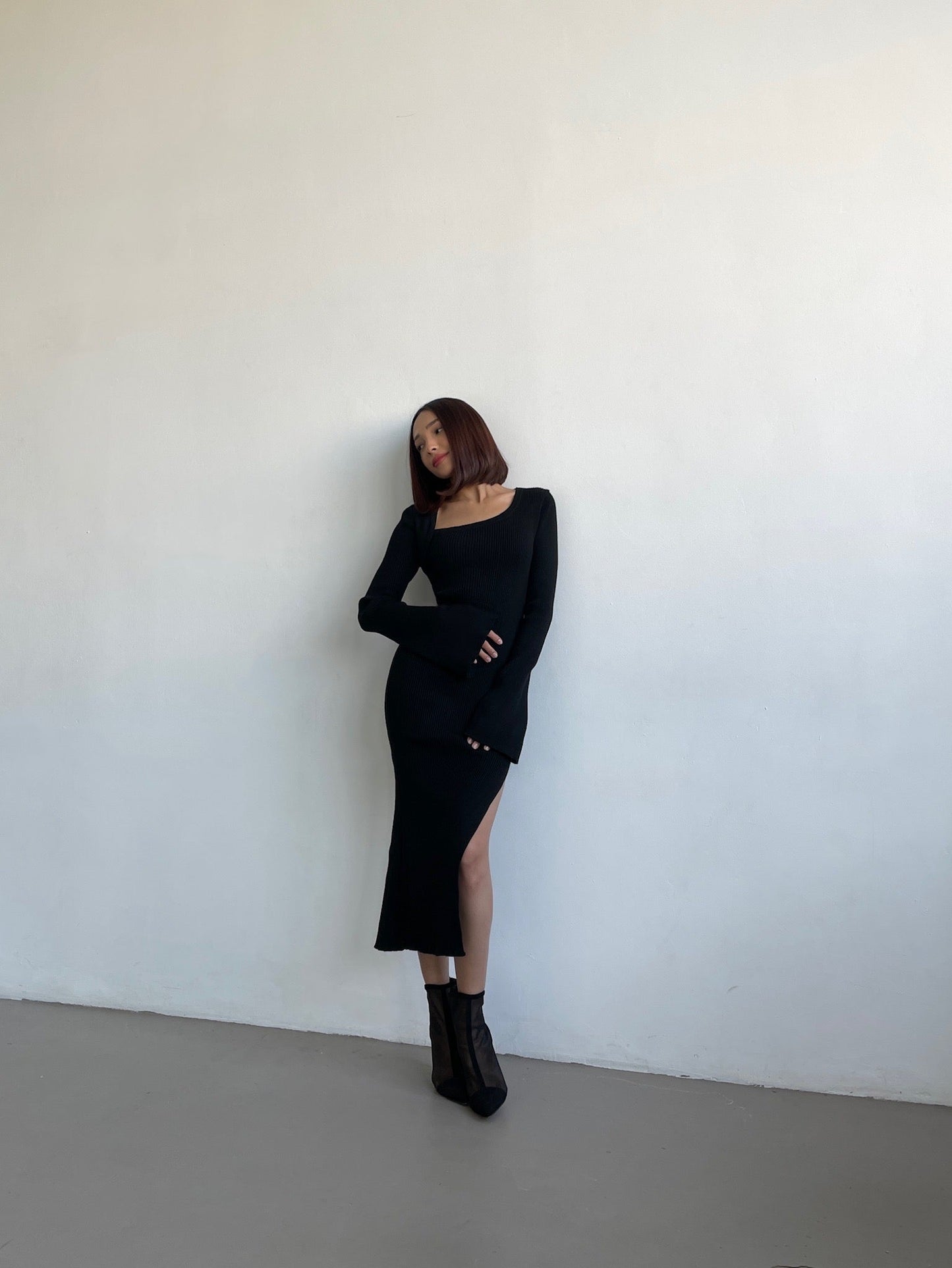 Against a plain white wall, someone with shoulder-length hair wears the Anja knit dress—a form-fitting black midi dress with long sleeves and a high slit—paired with black ankle boots. Their left hand rests on their hip, while their right arm crosses their body.
