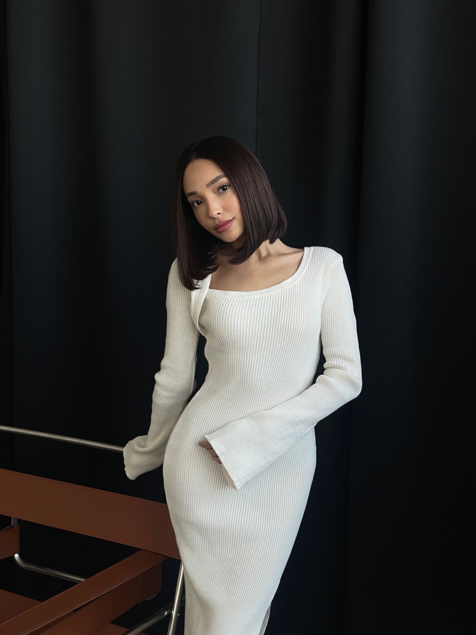 A person with shoulder-length dark hair wears the Anja knit dress, a white, long-sleeved midi dress featuring flared cuffs and a high slit. Leaning slightly against a railing in front of a dark background, they exude a minimalist and elegant ambiance.
