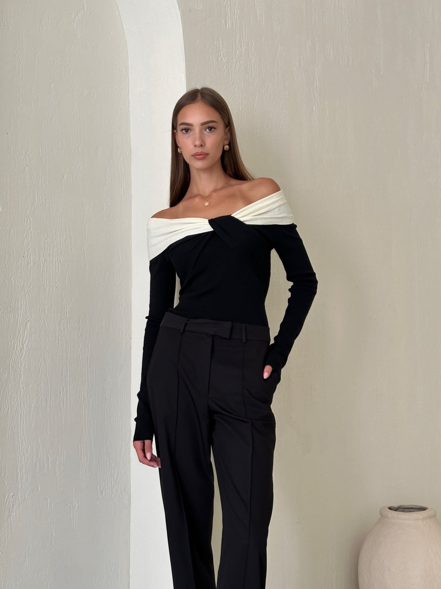 A woman stands against a light-colored wall wearing the stylish Amélie knit top, featuring an off-the-shoulder design with a contrasting white wrap detail, paired with black trousers. She has long, straight hair and poses with one hand in her pocket, exuding a sophisticated look. An urn-shaped vase is visible on the floor to her right.
