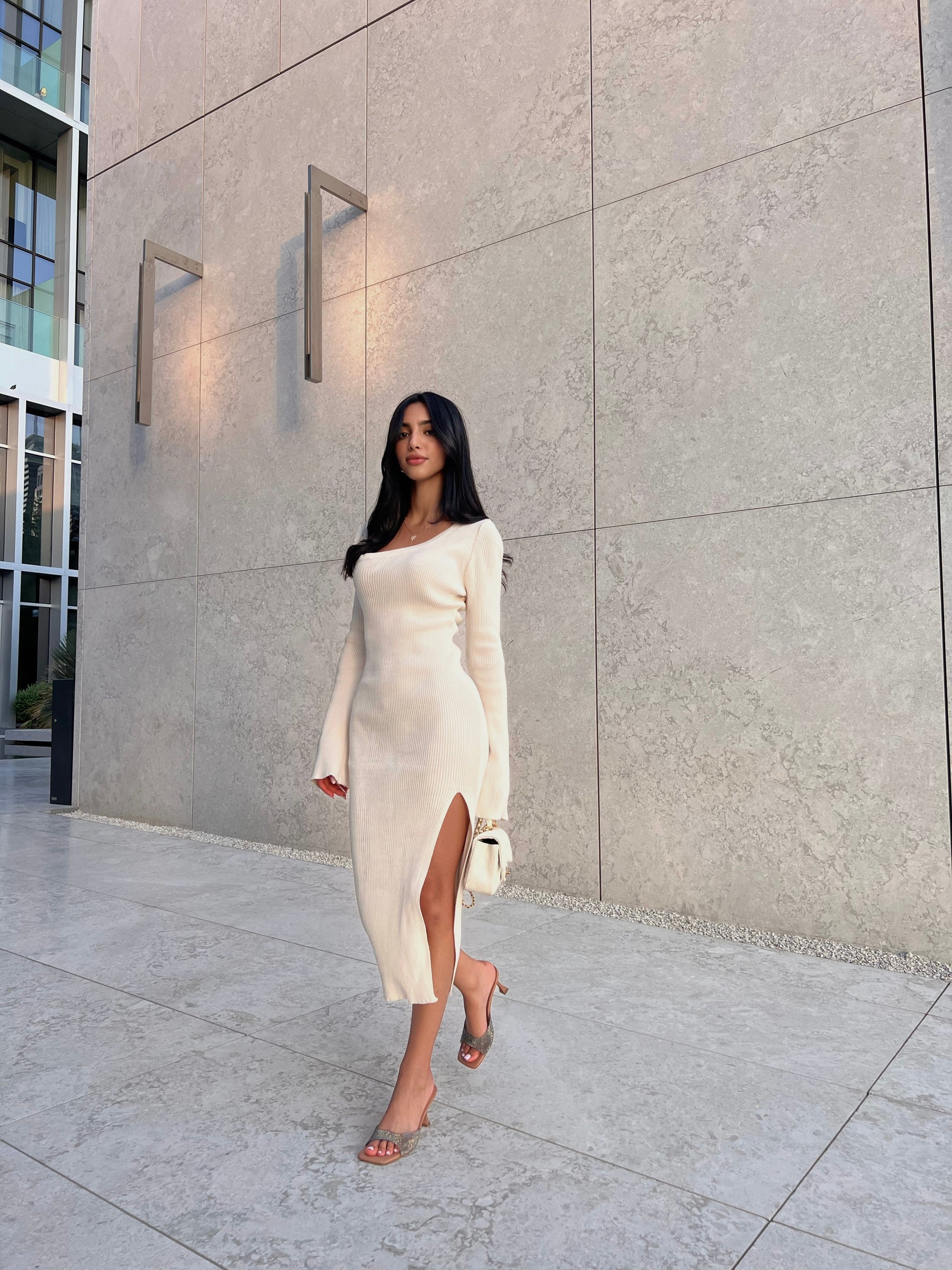 A woman with long black hair confidently walks along a tiled sidewalk in the form-fitting Anja knit dress, featuring a high slit. She carries a small white handbag, set against a backdrop of a modern beige stone wall with sleek lighting fixtures.