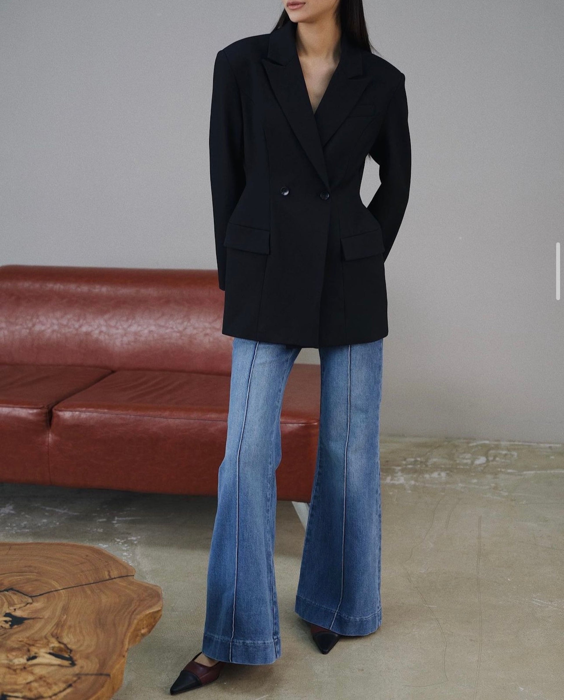 A person with long dark hair stands confidently in a black Olivia tailored blazer and wide-leg blue jeans, posing in front of a brown leather sofa on a concrete floor, with the corner of a wooden table peeking into view.