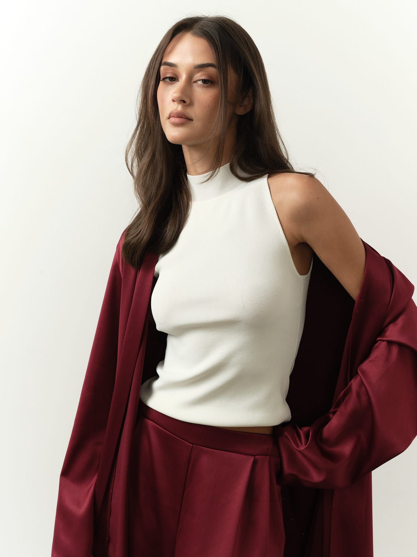 A woman with long brown hair confidently wears a sleeveless Maia Top underneath a draped burgundy jacket made from luxurious fabric, set against a neutral background, exuding sophisticated style.