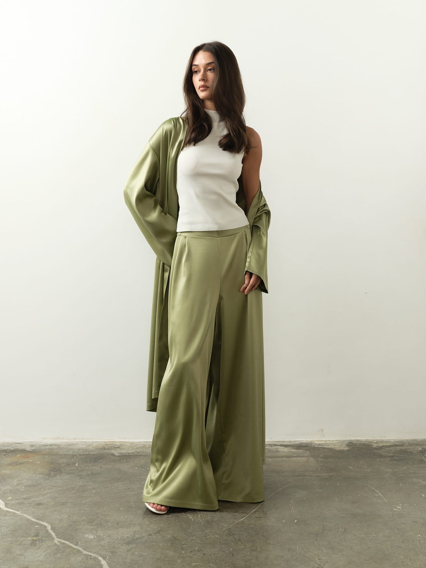 Against a plain white background, a woman presents her sophisticated style in a light green satin pantsuit and flowing robe. Her long dark hair complements the sleeveless Maia Top she wears as she strikes a pose with one hand in her pocket.