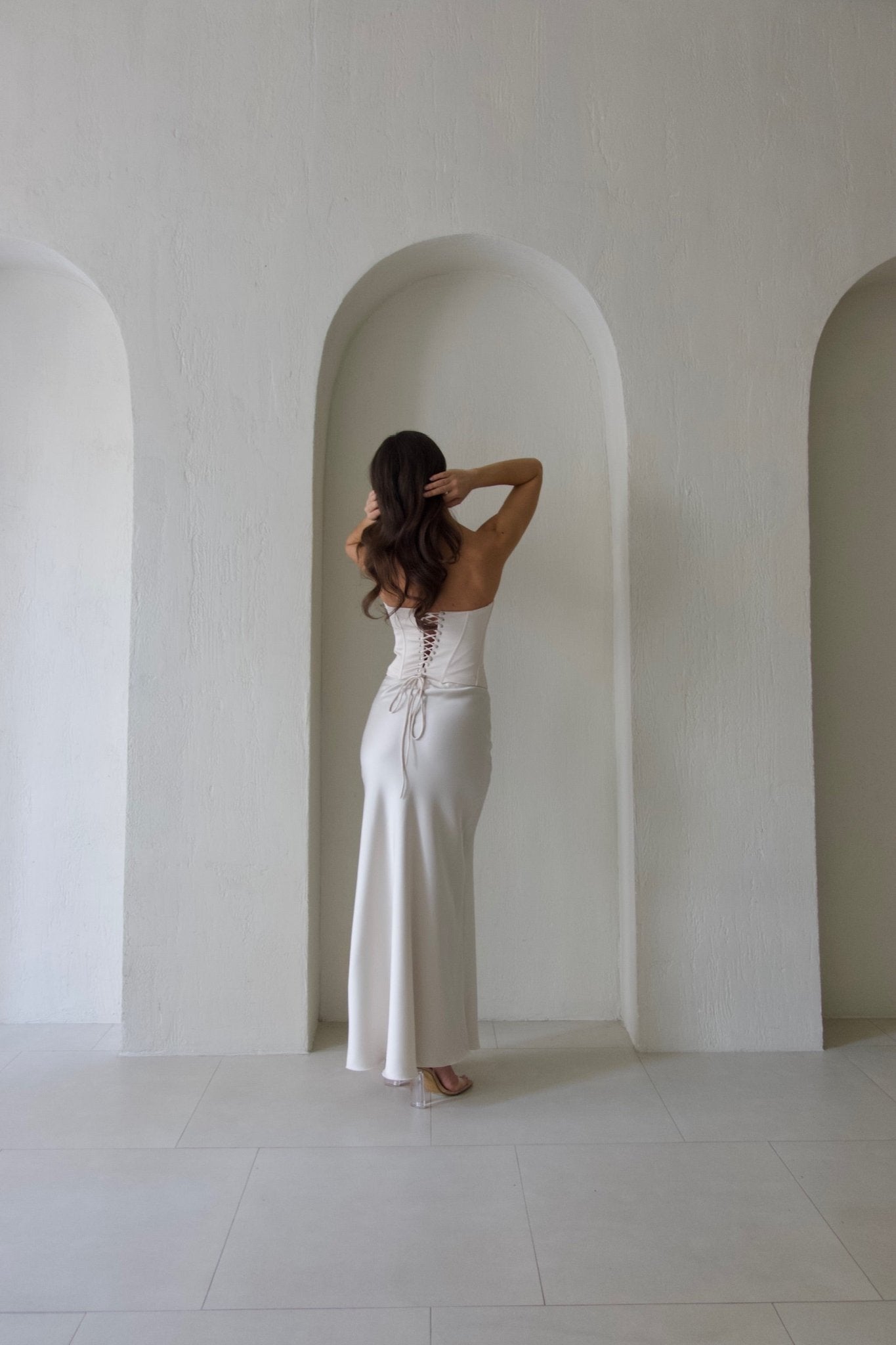 A woman with long, wavy hair stands with her back to the camera, adjusting her hair with both hands. She is wearing the Regina corset set with a maxi skirt in a form-fitting, strapless white design made of satin fabric and featuring a lace-up back. She is standing in front of a plain, light-colored wall featuring a recessed arch.
