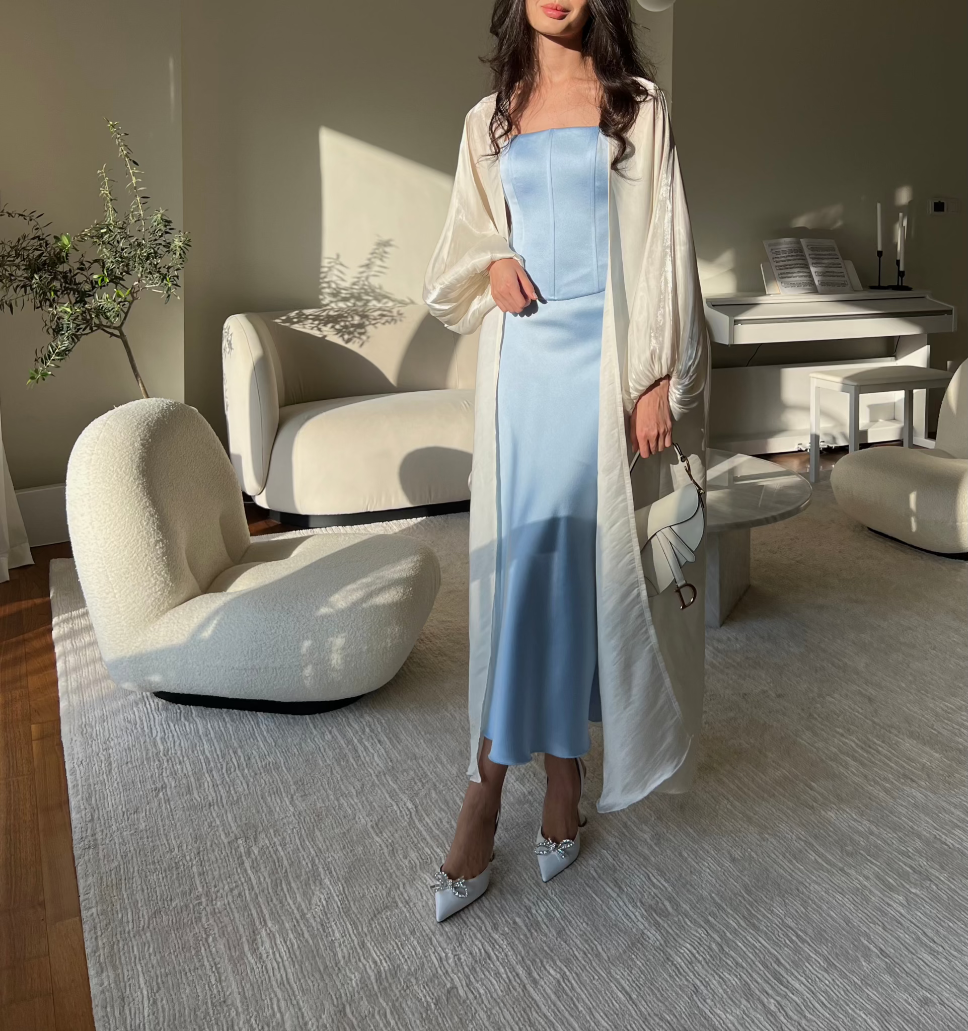 A person stands in a modern, sunlit living room wearing a Regina corset top paired with a light blue dress with a white sheer overlay and white pointed heels featuring a bow detail. The room, adorned with elegant white furniture including a chair, sofa, and piano, exudes sophistication reminiscent of Bridgerton-esque corsets, with a plant adding a touch of greenery.