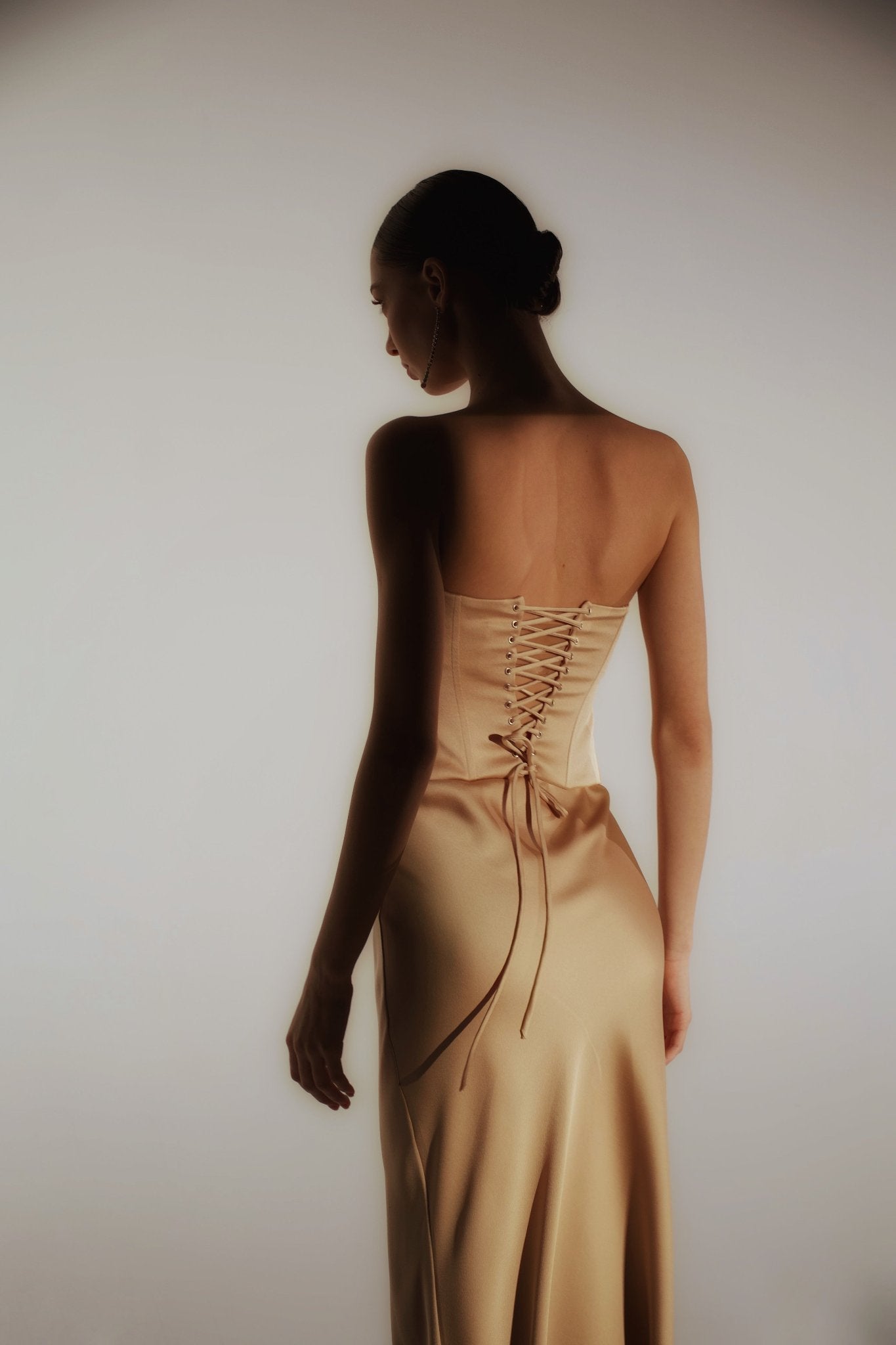 A woman with dark hair styled in a bun is shown from behind, wearing the Regina corset top in satin, styled with lace-up and strapless detailing in a soft, neutral shade. The image is softly lit, highlighting the elegant drape and sheen of the sculpting satin fabric. The background features a gradient light tone.